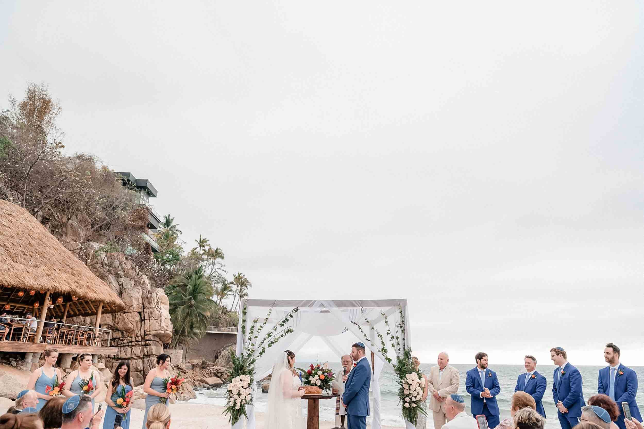 Wedding ceremony on the beach with bride and groom standing under an arch, surrounded by wedding party and guests, with ocean and sky in the background.