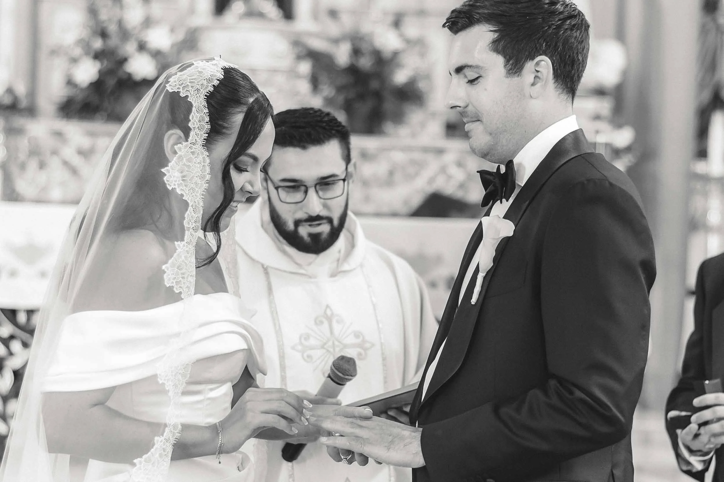 A wedding ceremony with a bride and groom exchanging vows, with an officiant holding the Bible, in a church with floral decorations in the background.