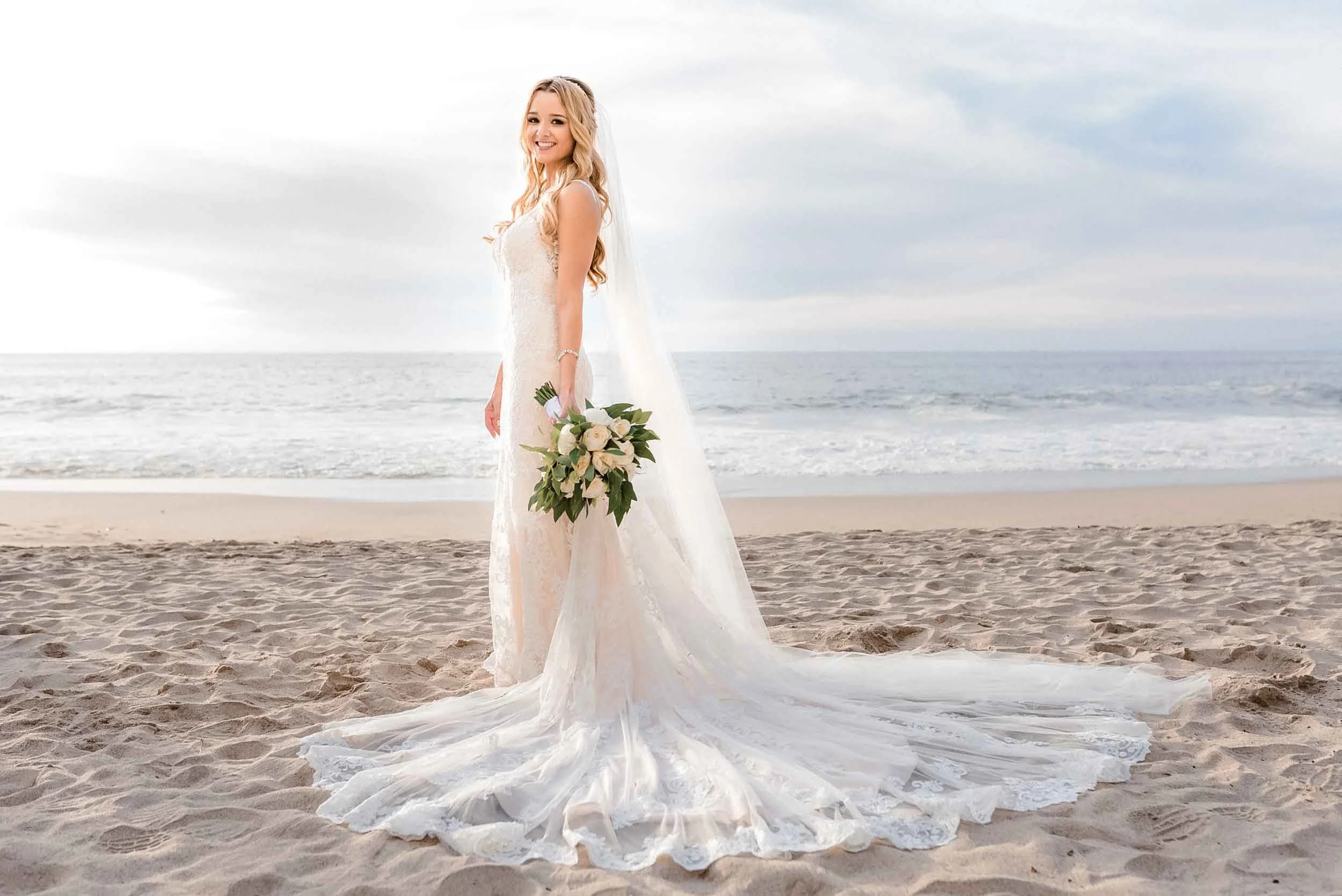 A smiling bride in a white wedding dress holding a bouquet of white roses and greenery, standing on a sandy beach with the ocean and a cloudy sky in the background.