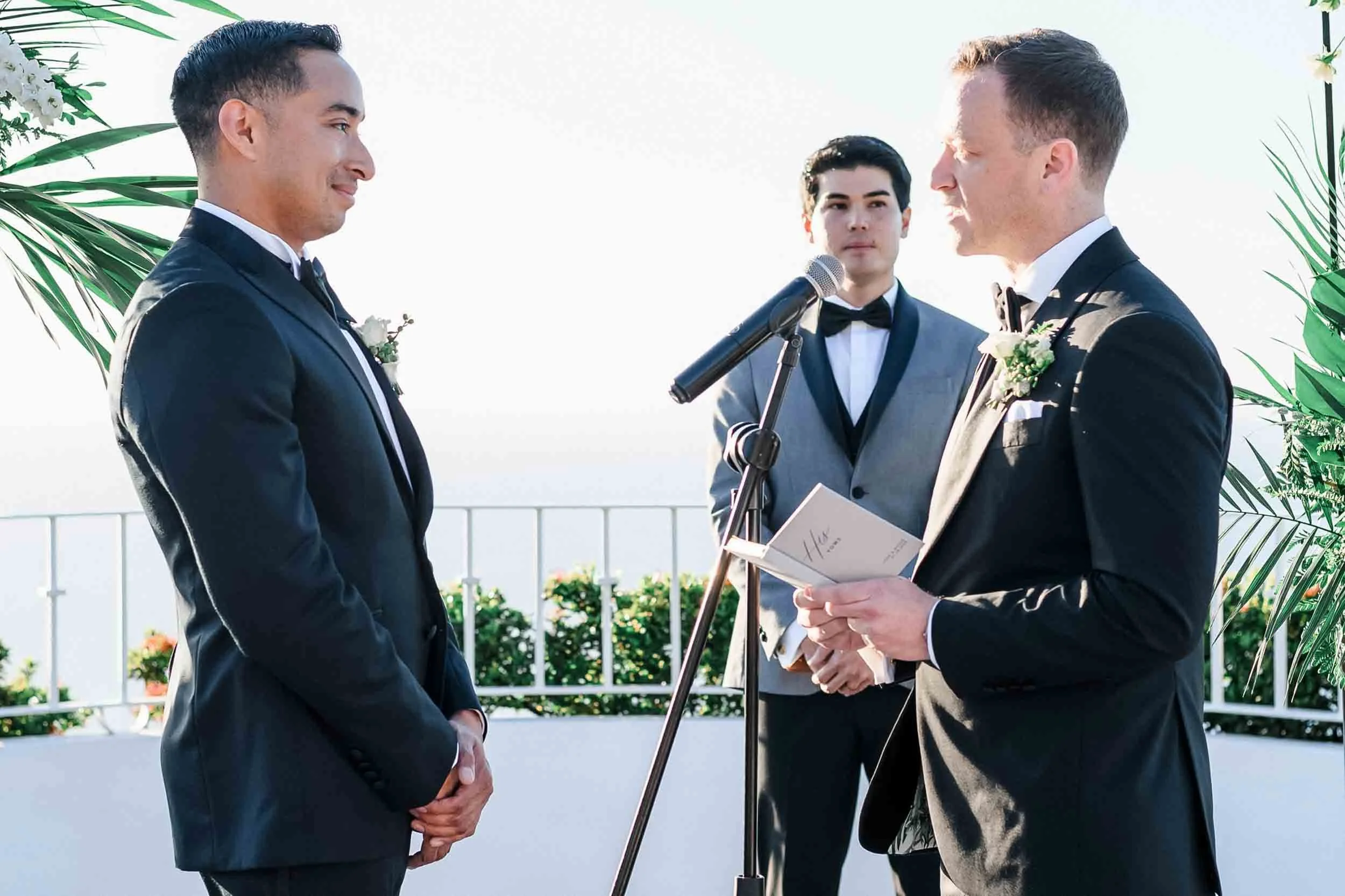 A LGBTQ couple getting married outdoors, exchanging vows, with an officiant reading from a book, greenery, and ocean in the background.