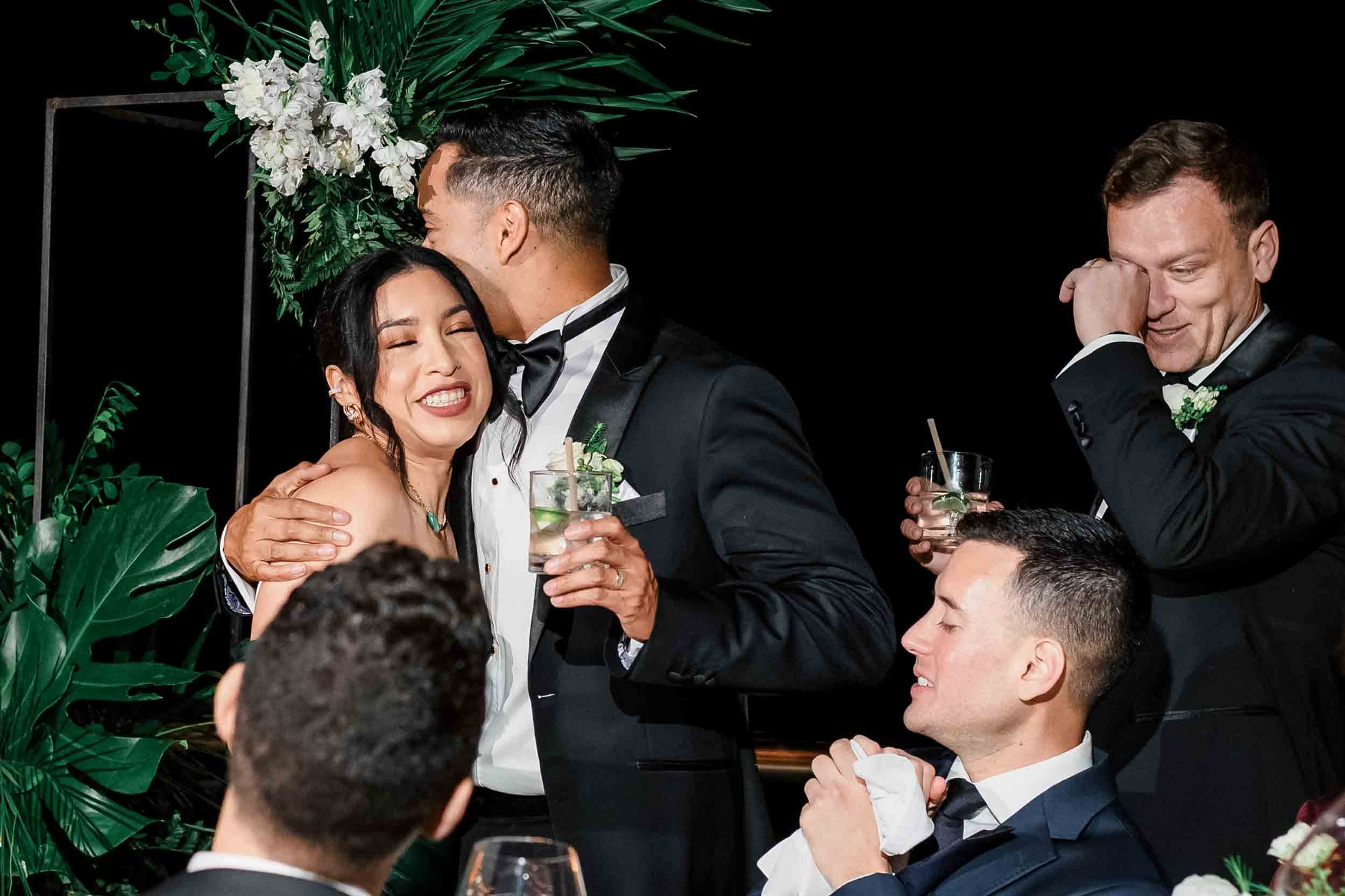 Grooms sharing an emotional moment at their wedding reception, with a background of lush green tropical leaves and white flowers.