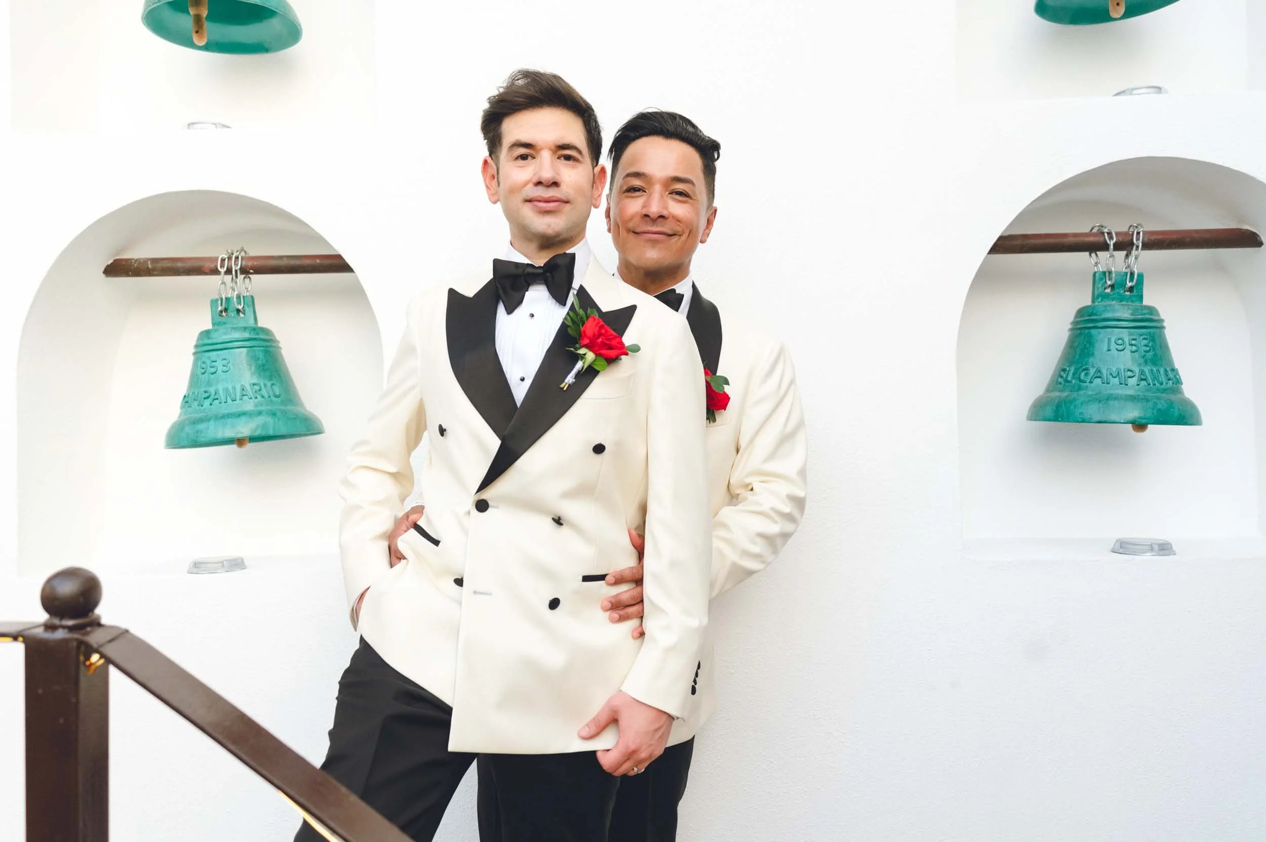 Grooms in tuxedos posing together during portrait time after they lgbtq wedding,  standing in front of white walls with green bells hanging on the walls. 