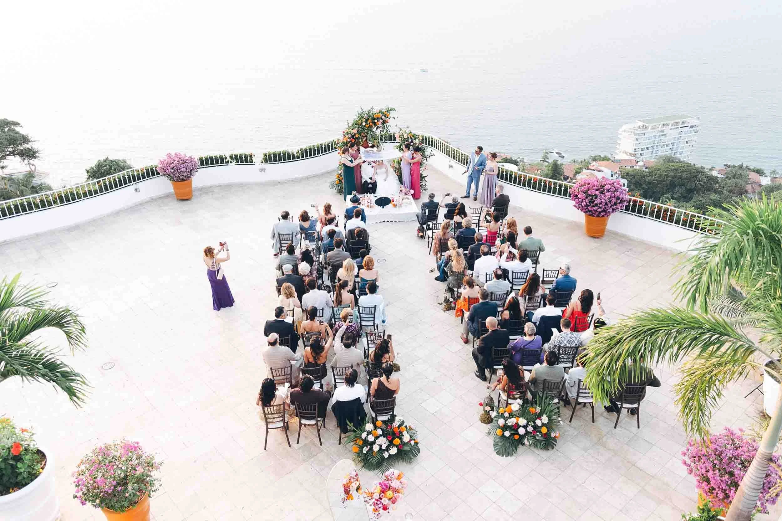 A wedding ceremony taking place on a rooftop terrace overlooking the water in Puerto Vallarta, with guests seated and the bride and groom standing at the altar surrounded by floral arrangements.
