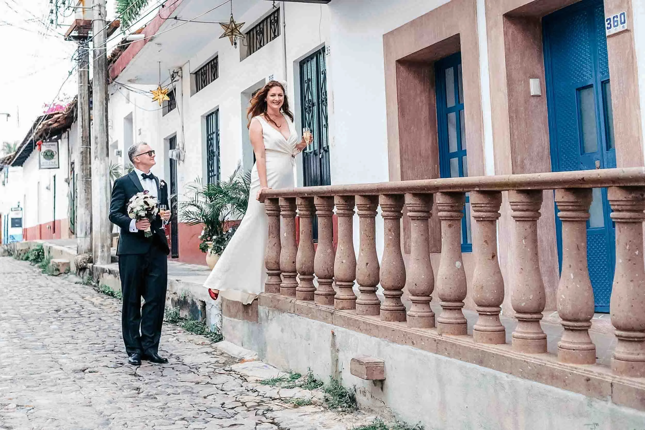 Bride standing on sidewalk , holding a glass of champagne, smiling. Groom with her bouquet and a glass of champagne in his hand stands on cobblestone street looking up at her. The scene is outdoors in the colorful town of Vallarta with tiled rooftops