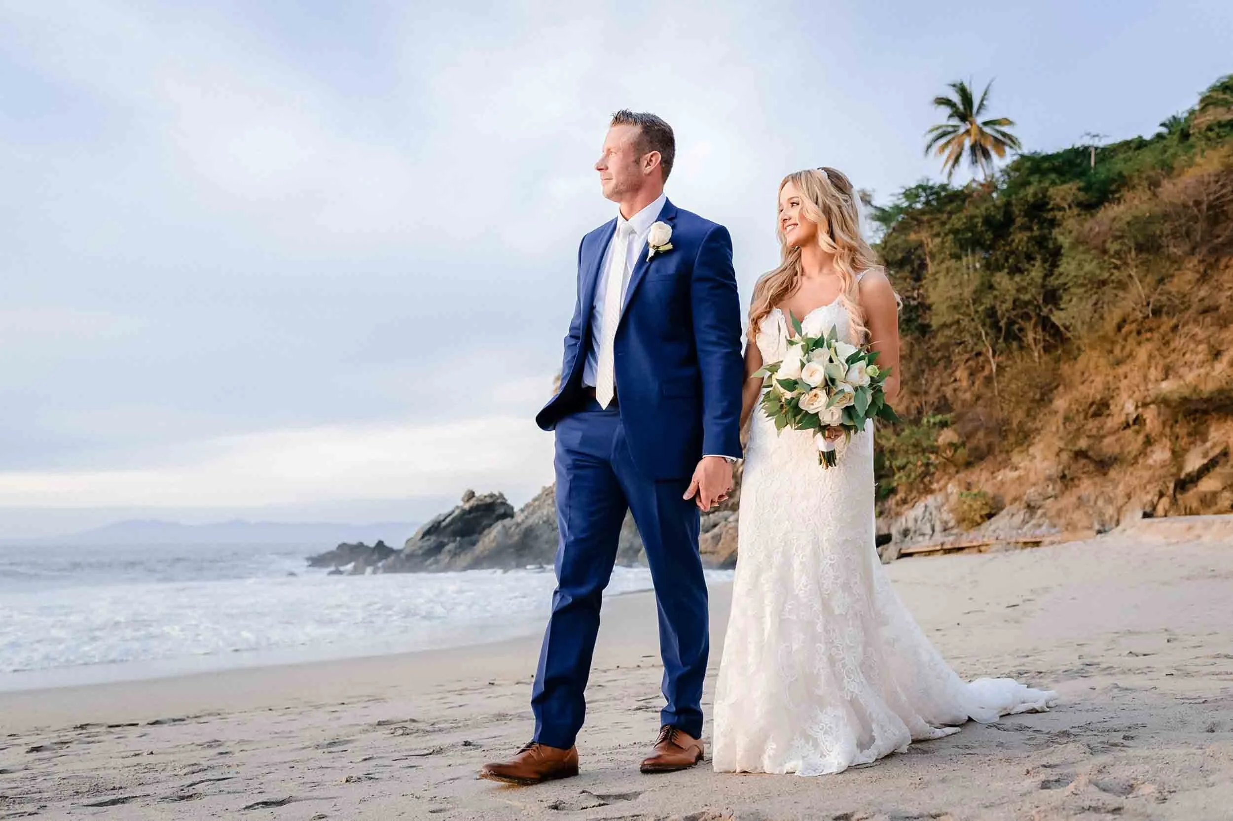 A bride and groom holding hands on a beach, with the ocean and rocky cliffs in the background at the Hyatt Ziva in Puerto Vallarta.