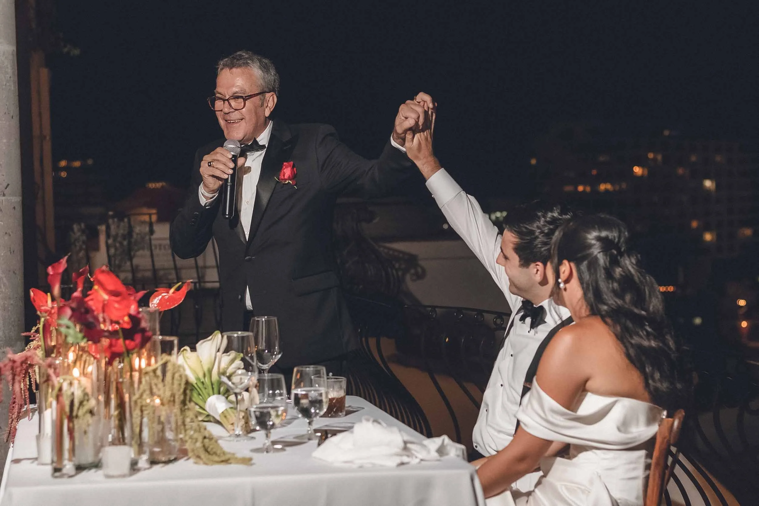 Father of the bride  giving a speech at the wedding reception on a rooftop at night with city lights in the background.