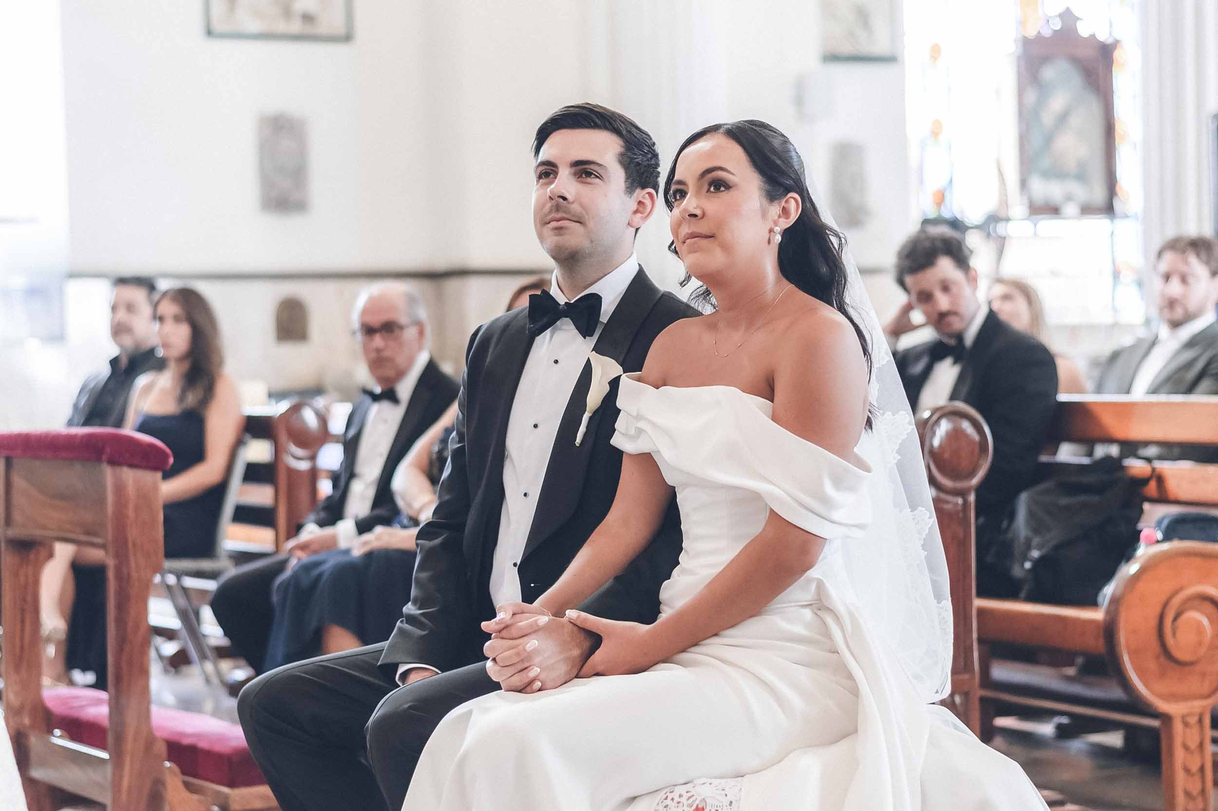 A bride and groom sitting in a church during their wedding ceremony, surrounded by guests.