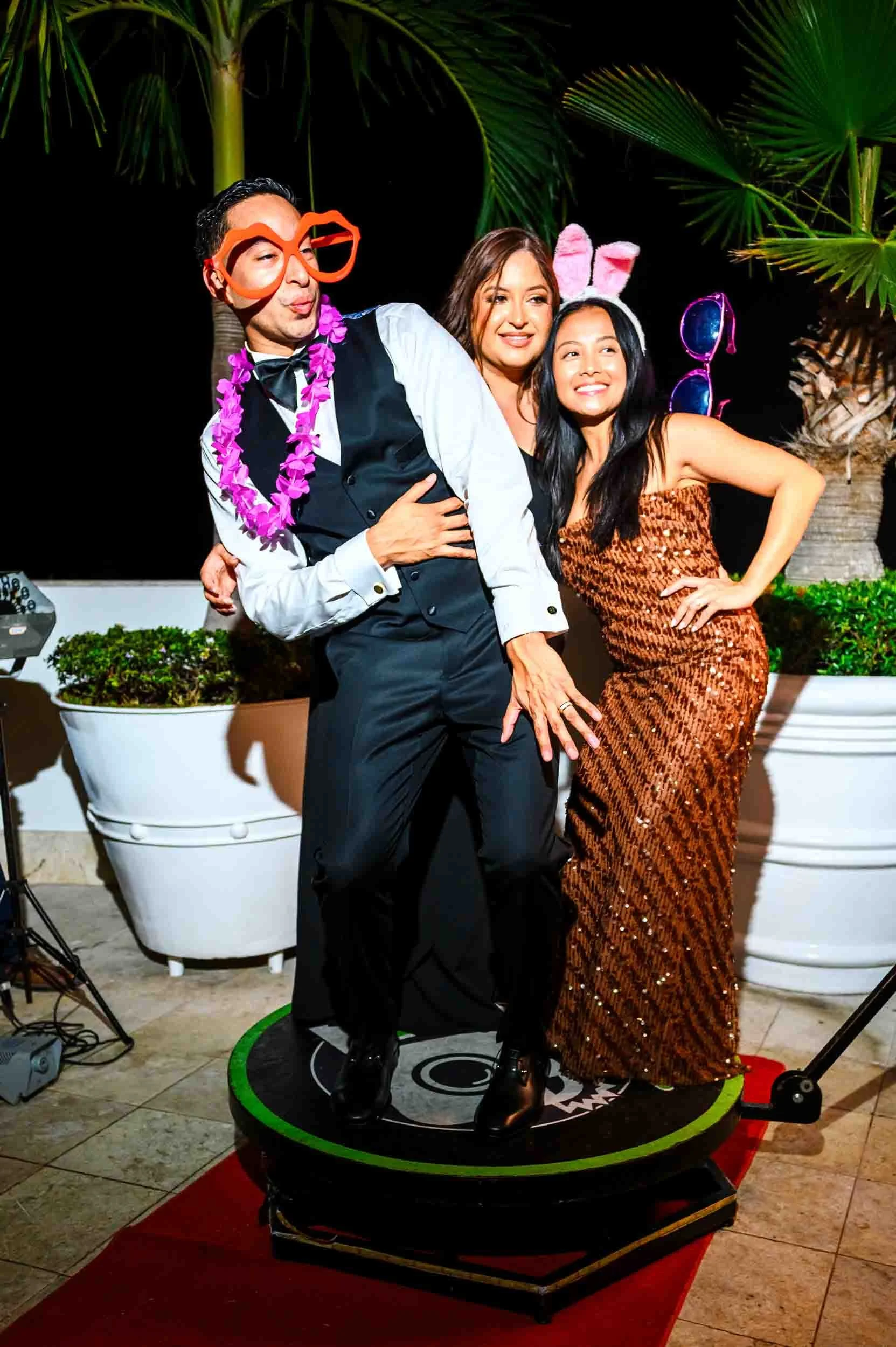 Three people at a party posing on a small platform and posing for photos during a wedding reception, the one with the big glasses is the groom. 
