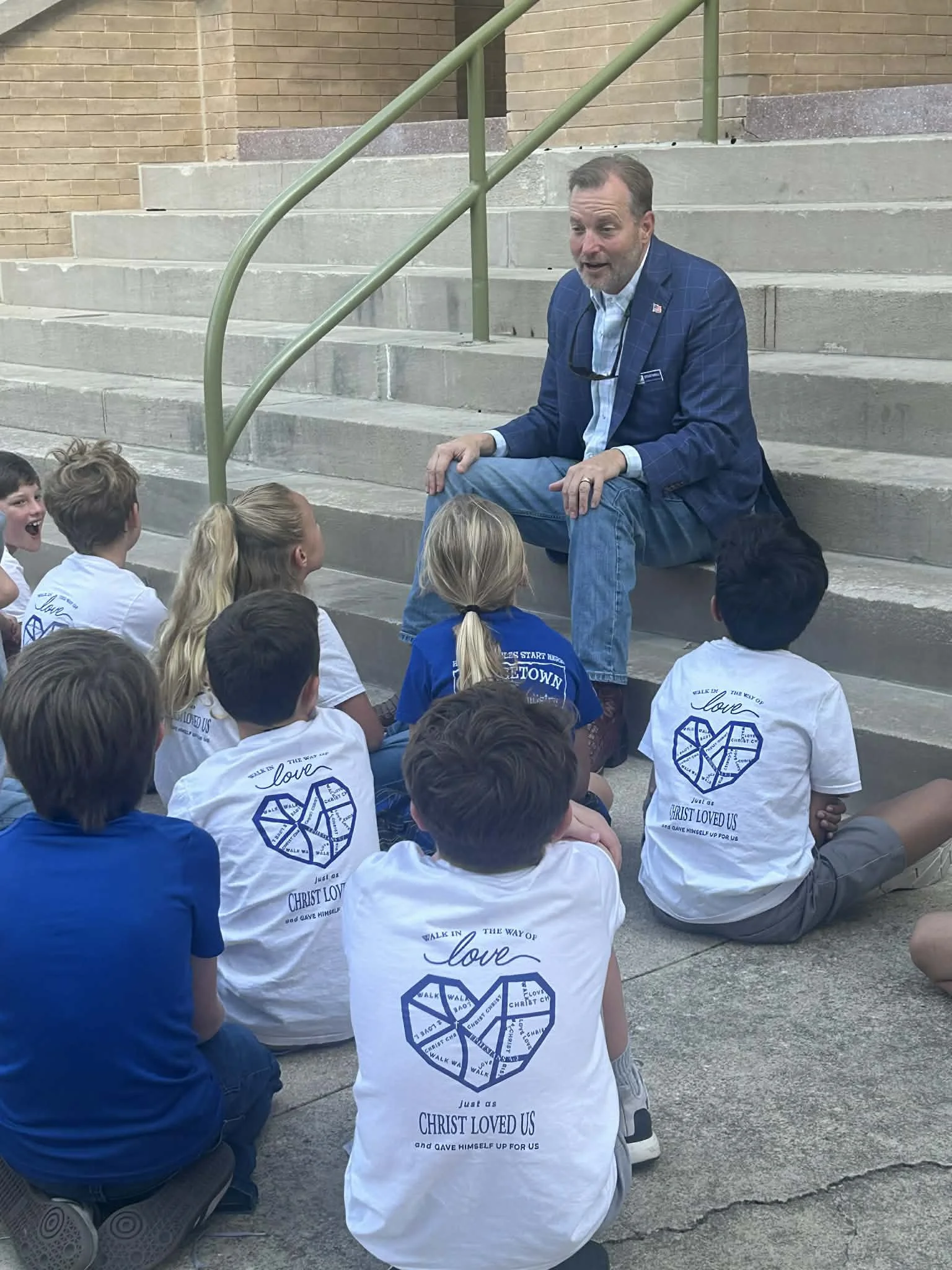 A man in a blue blazer sitting on concrete stairs talking to a group of children. The children are kneeling or sitting and wearing white shirts with a heart-shaped design and text about Christ's love.
