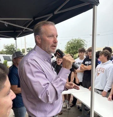 A man with gray hair and beard, wearing a purple dress shirt, speaking into a microphone outdoors under a black canopy, with a group of young men listening.