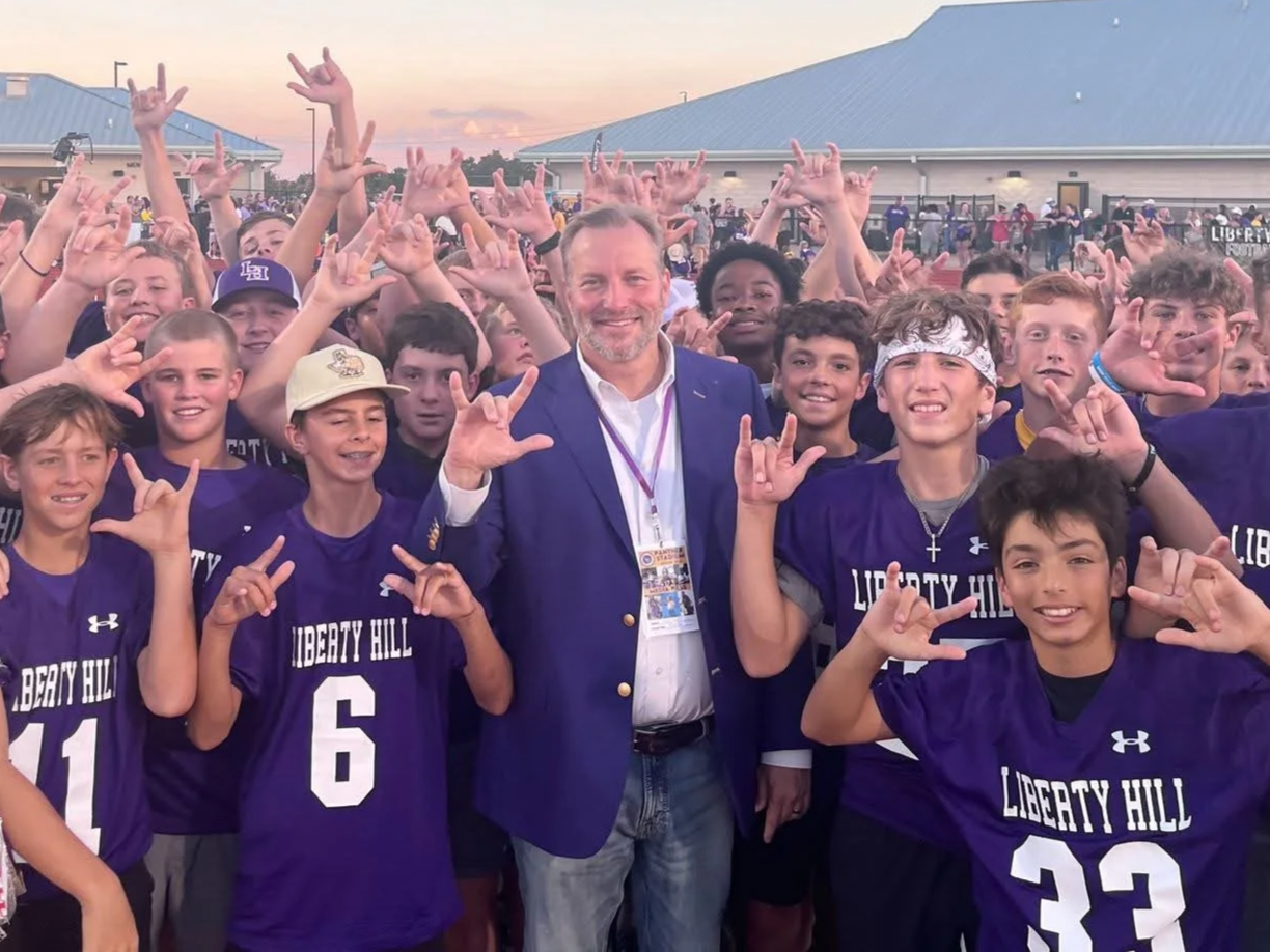 Group of young football players in purple jerseys with 'Liberty Hill' written on them, posing with a smiling adult male coach or organizer, making hand signs, outdoors during sunset.
