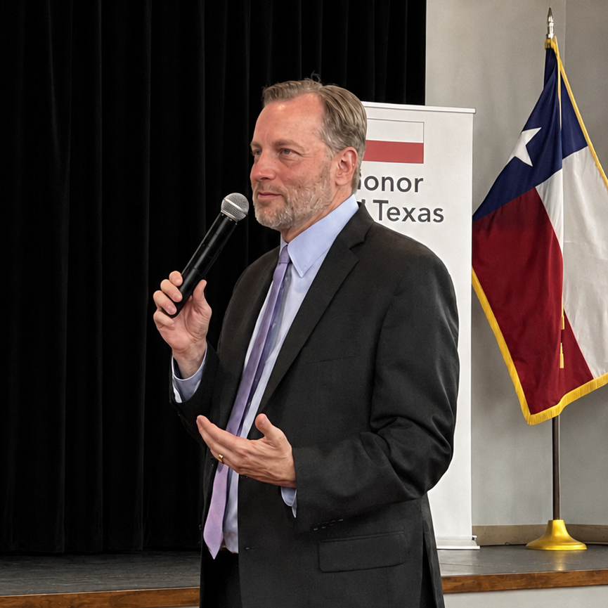 A man in a suit and tie speaking into a microphone during an event. Behind him is a Texas flag and a banner that reads 'Honoring Texas'.