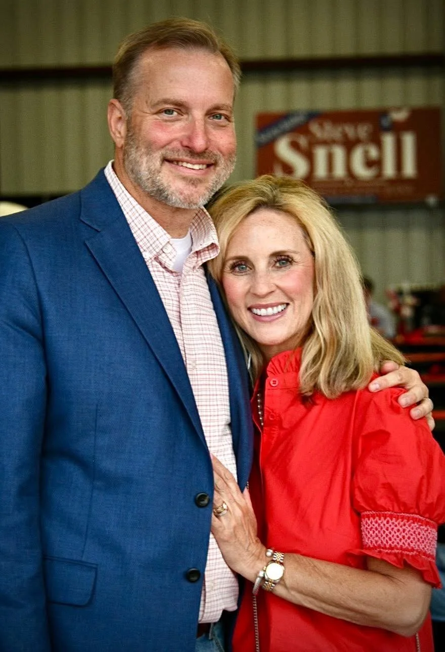 A smiling man and woman posing together indoors. The man is wearing a blue blazer and a checked shirt, and the woman is wearing a red blouse. They are embracing, and there is a blurred sign in the background.