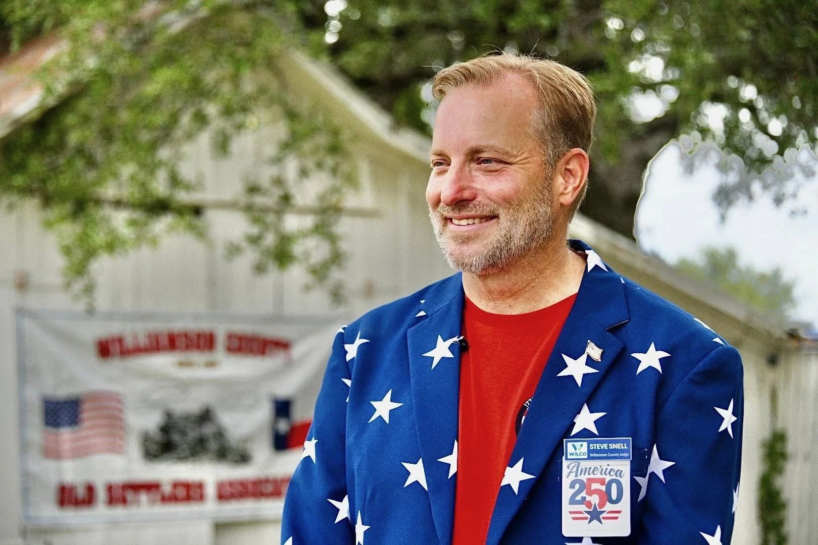 A smiling man in a patriotic star-spangled jacket and red shirt standing outdoors during daytime with trees and a banner in the background.