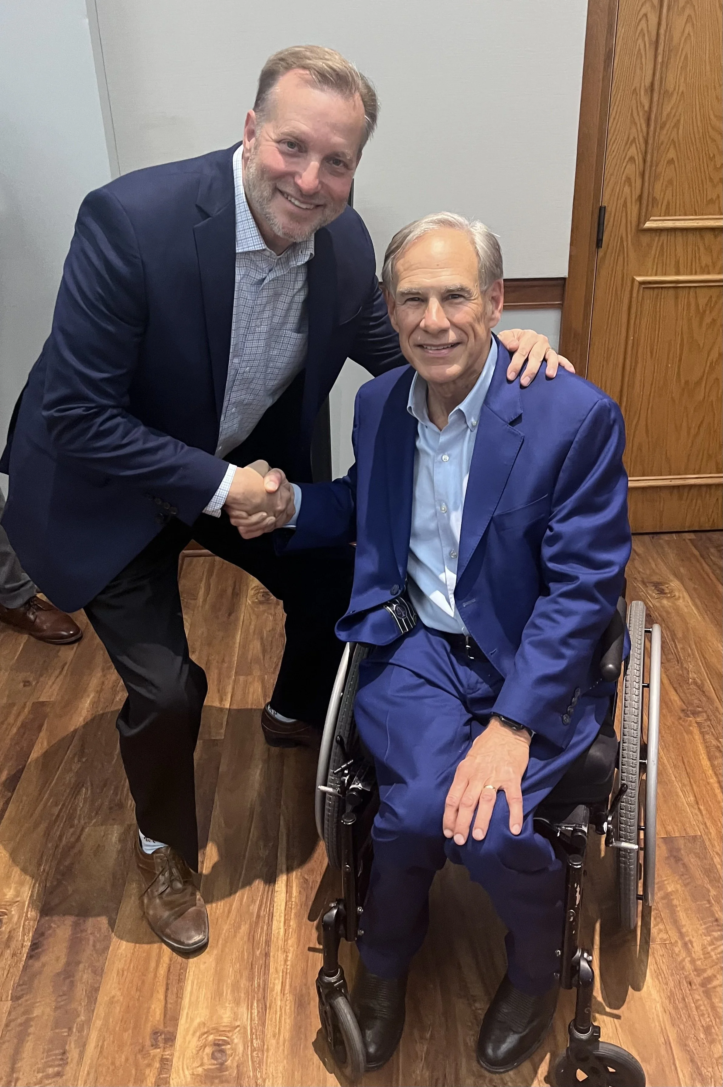 Two men, wearing blue suits, shaking hands. One man is sitting in a wheelchair, and the other is standing next to him. They are smiling and posing for the photo in an indoor setting with wood paneling and hardwood floor.
