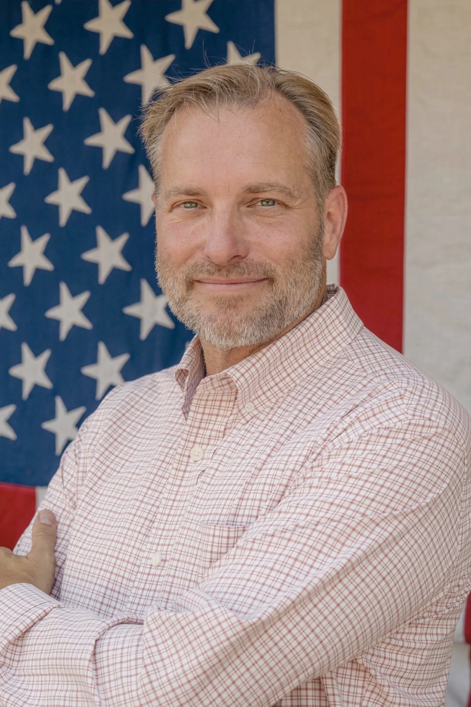 A man with light skin, gray beard, and blond hair standing in front of an American flag, wearing a light pink, checked dress shirt.