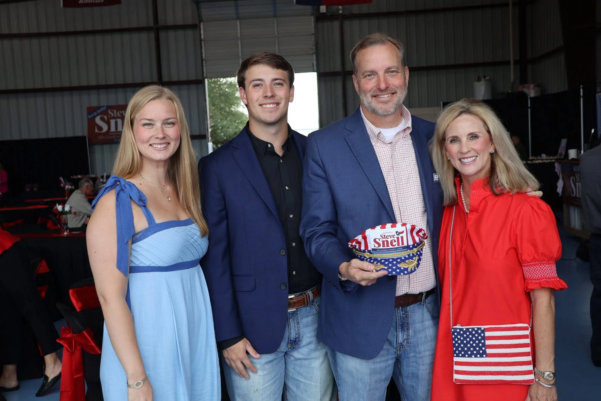A group of four people standing together inside a building, smiling for the photo. One man in a blue blazer is holding a decorated cowboy hat with patriotic colors and the name "Steve Snell." The women are dressed in casual summer clothing, and the man is wearing a collared shirt. The background shows tables with items and a sign that reads "Steve Snell."