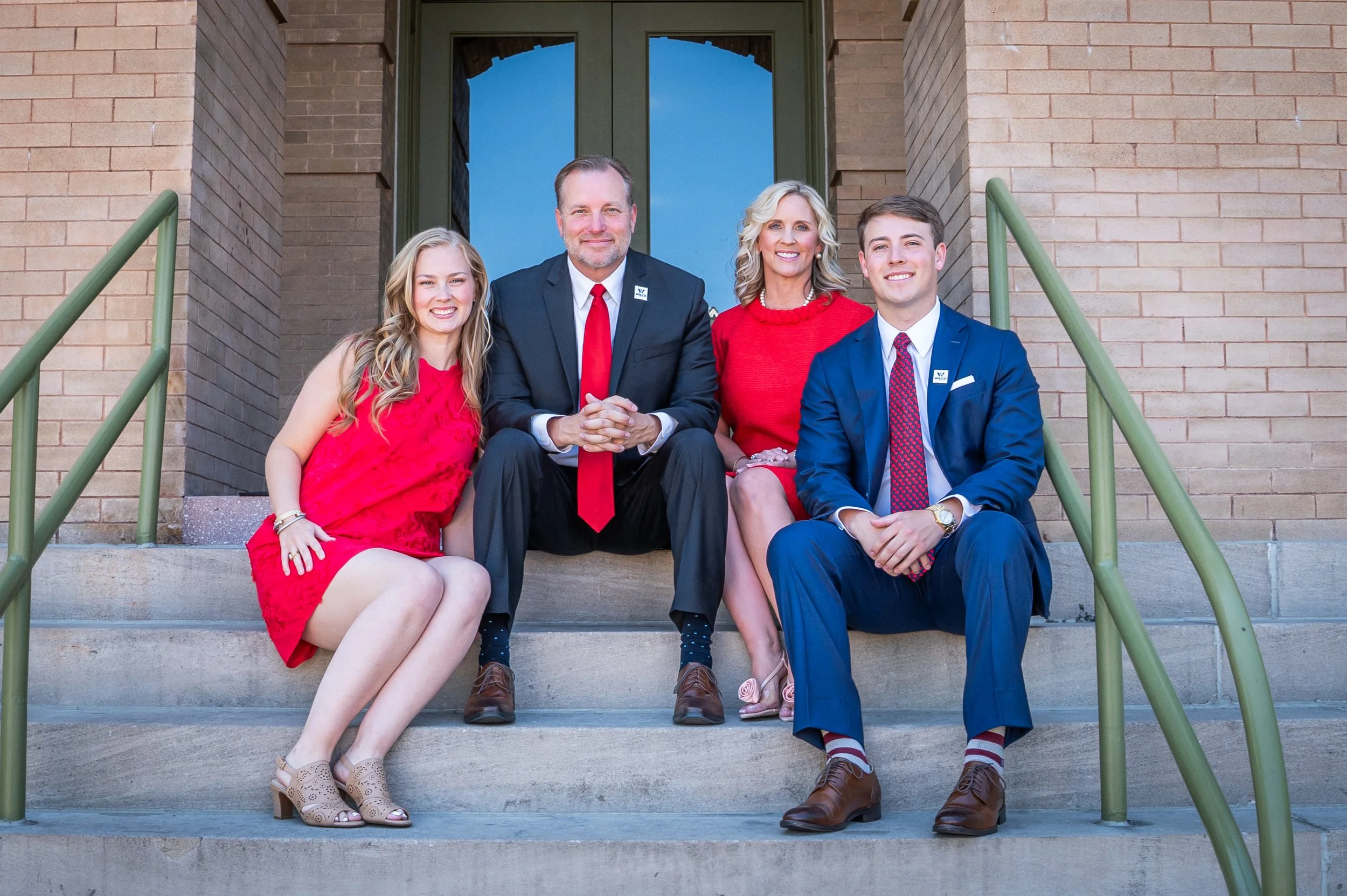 Family of four sitting on front steps of a brick house, smiling at the camera. The parents are in the middle, with the daughter on the left in a red dress and the son on the right in a blue suit. All are dressed semi-formally.