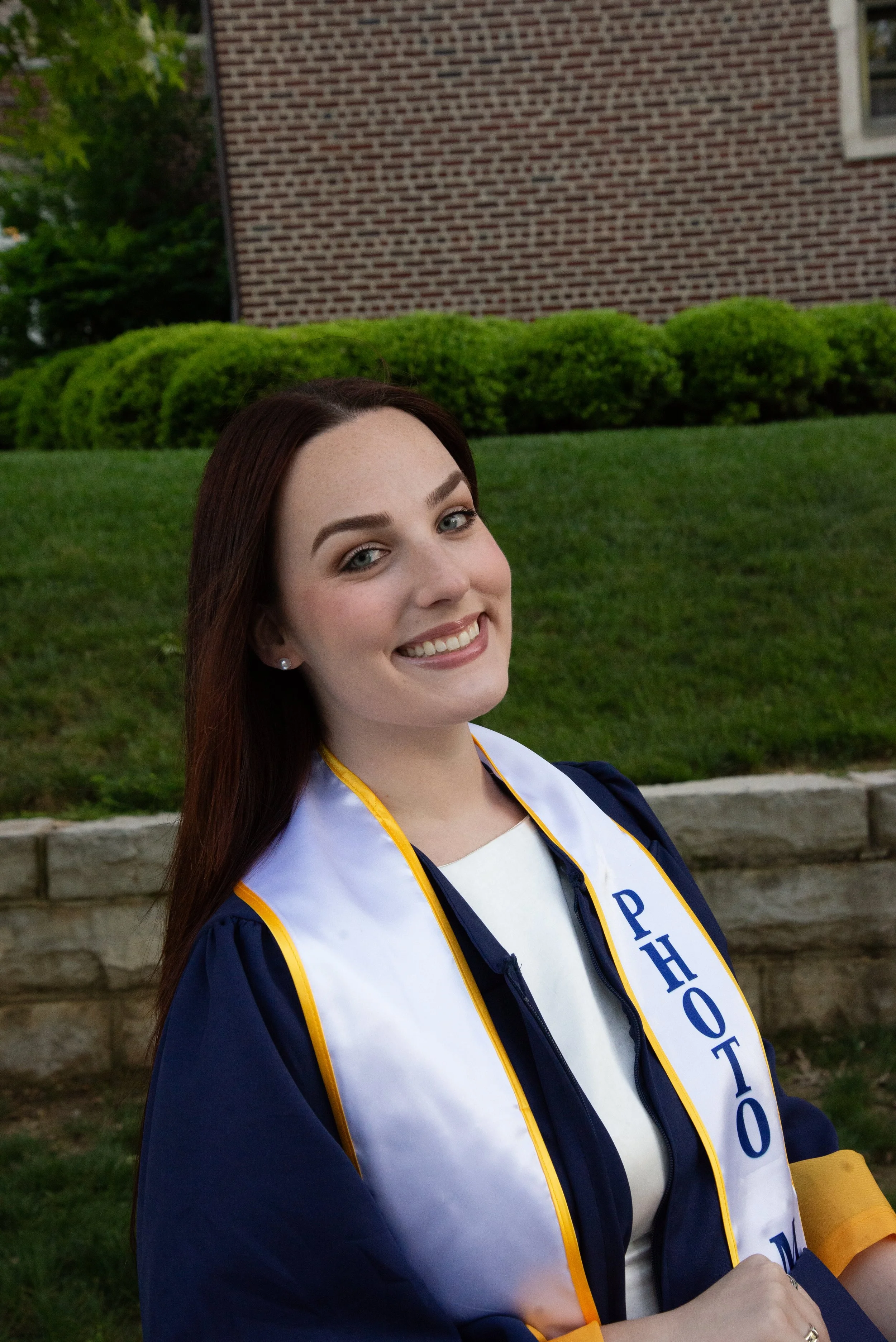 A young woman with dark red hair and light skin, wearing a graduation gown and stole, smiling outdoors in front of a brick building and green bushes.