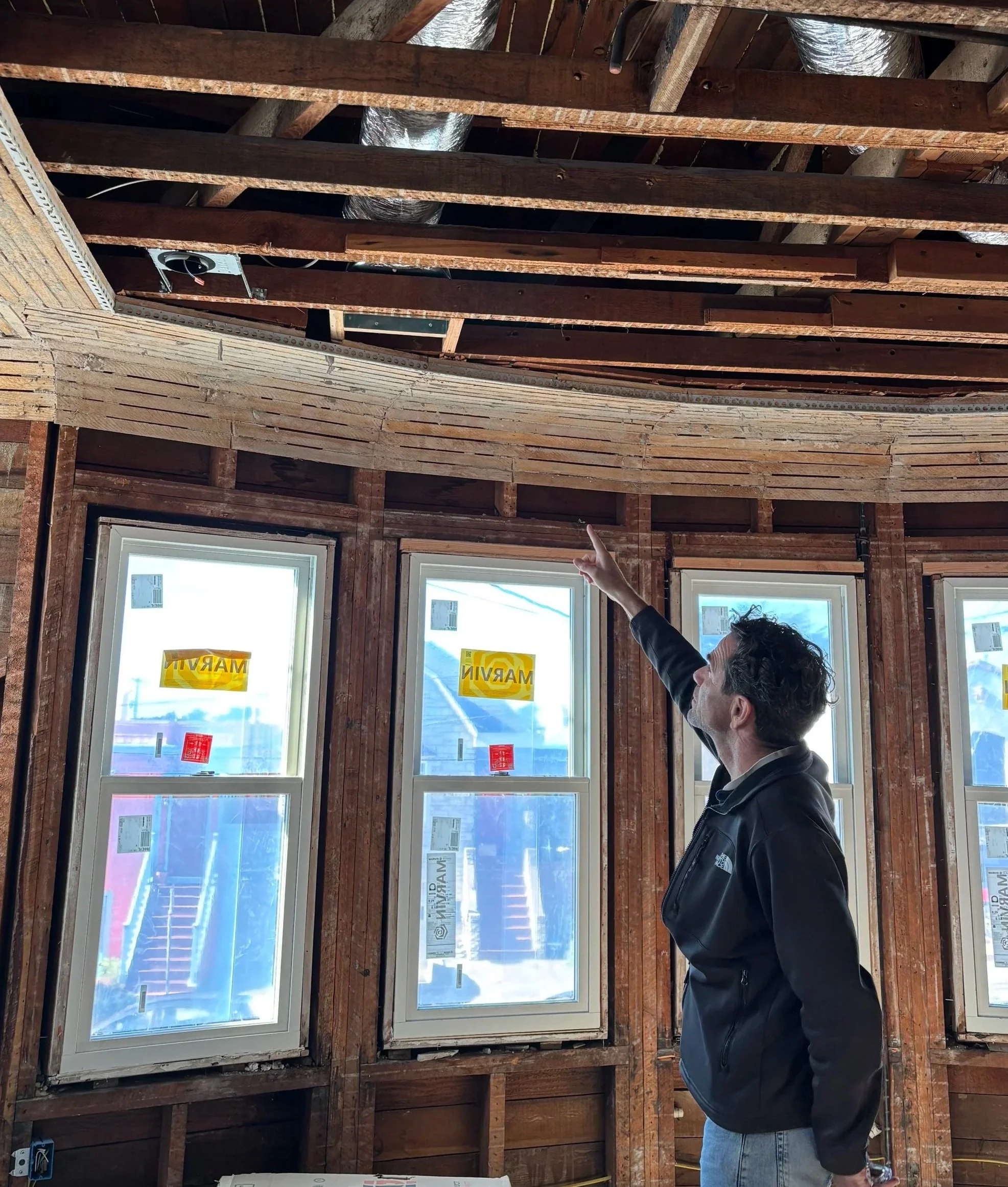 A person points towards the ceiling of a room under construction, showing exposed wooden framing and large windows with stickers on the glass. The person wears a black North Face jacket and blue jeans, and the room has visible electrical wiring and ductwork.