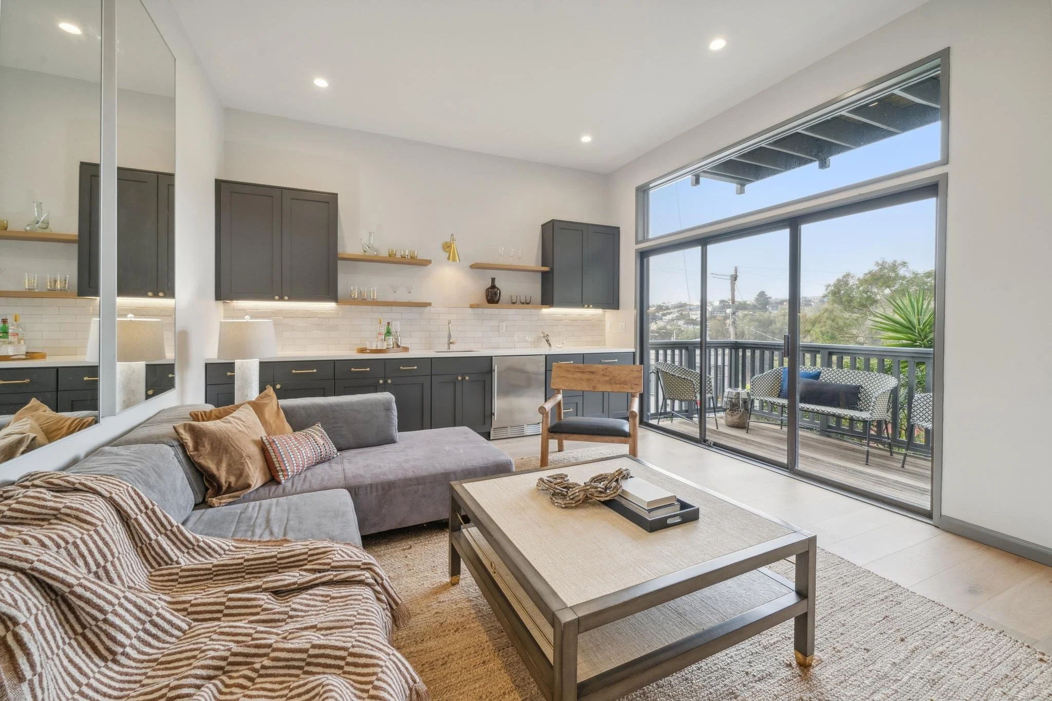 Living room with gray sofa, beige throw blanket, and pillows, coffee table with decorative items, large window and sliding door leading to a balcony with outdoor seating, kitchen area in background with gray cabinets, open shelves, and white subway tile backsplash.
