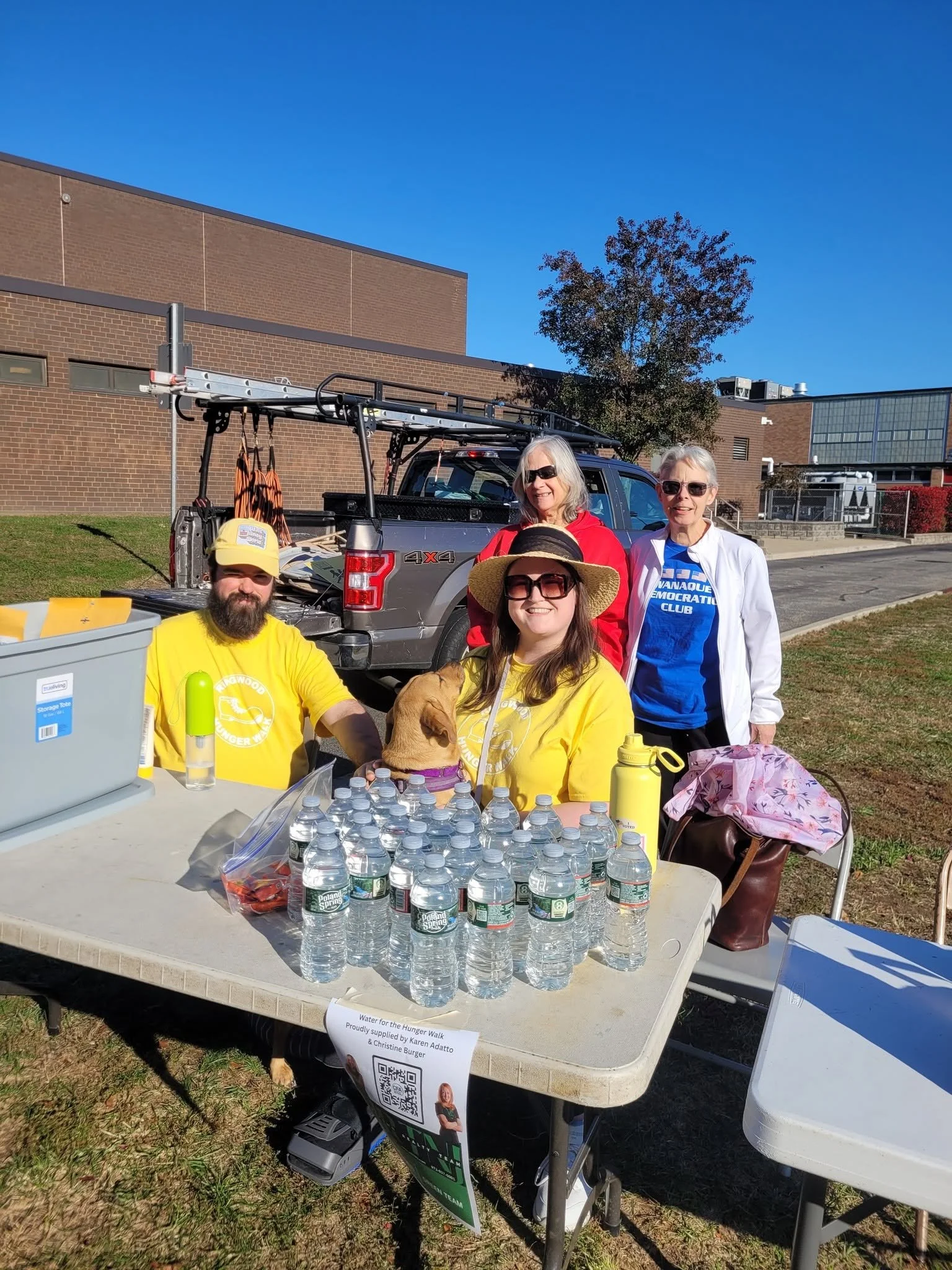A group of five people gathered outside at a table with bottled water and snacks. They are in front of a pickup truck with equipment in the bed, and behind them are brick buildings and a tree. One woman is wearing a white jacket and sunglasses, another woman is wearing a red jacket and sunglasses, and a man and woman are wearing yellow shirts with a dog logo. There is a sign on the table indicating the event is related to water for the Hunger Walk.