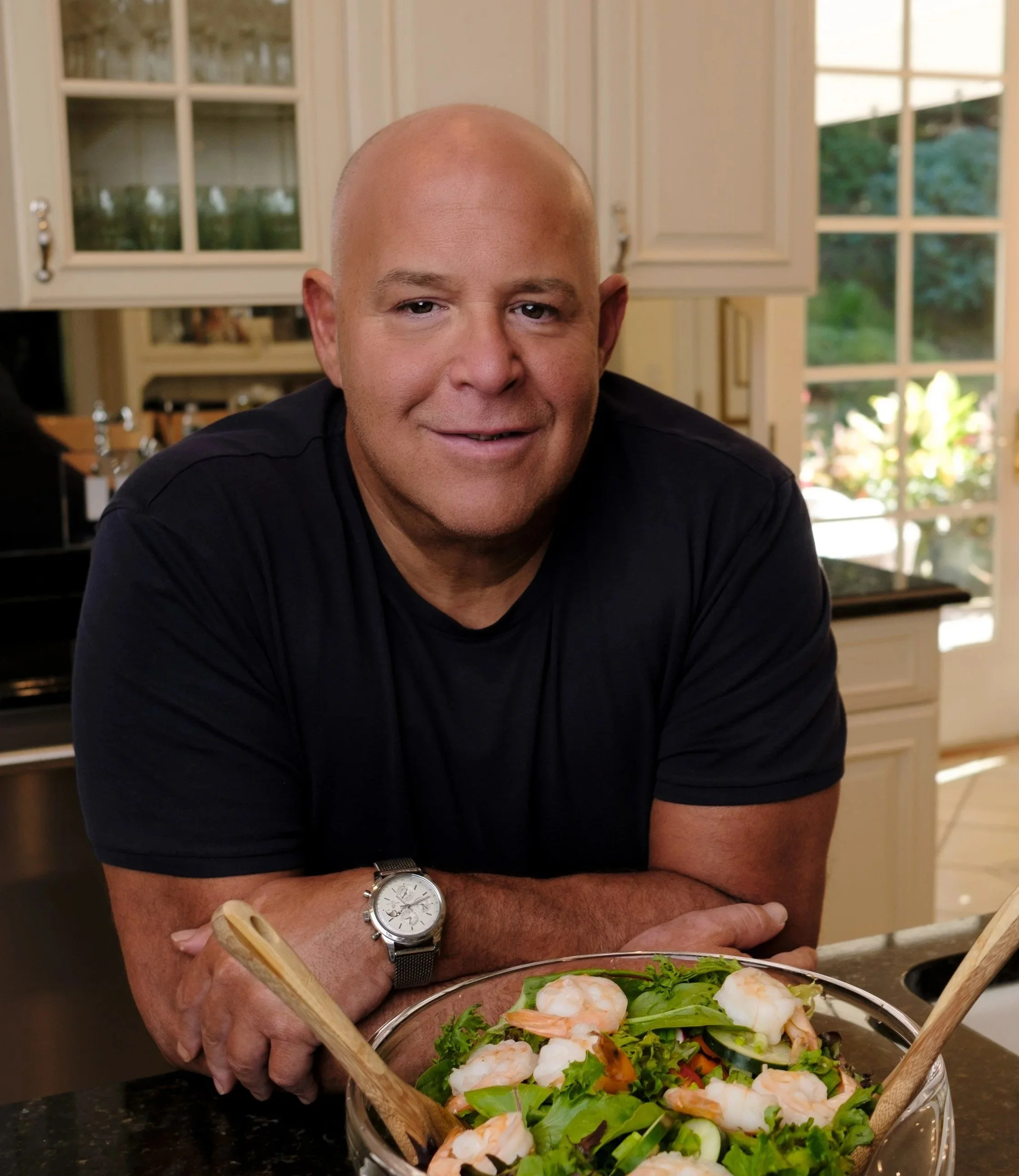 A man with a shaved head and a silver watch is preparing to serve a large salad with shrimp and vegetables in a kitchen with cream-colored cabinets and windows showing greenery outside.