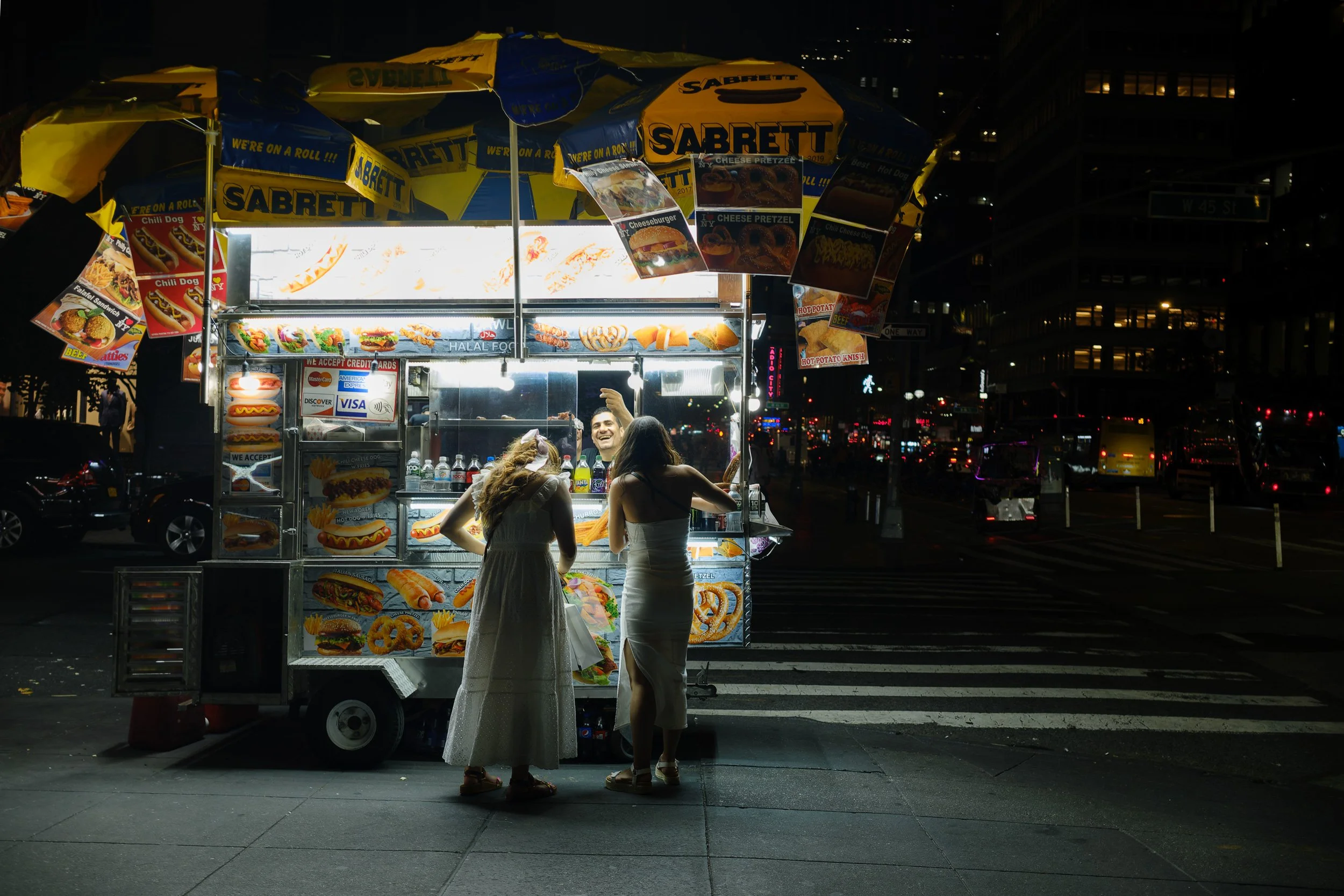 Night scene with two women talking at a hot dog stand decorated with colorful signs and umbrellas with the Sabrett logo. The stand displays picture menus of hot dogs, fries, and onion rings, with support for credit card payments.