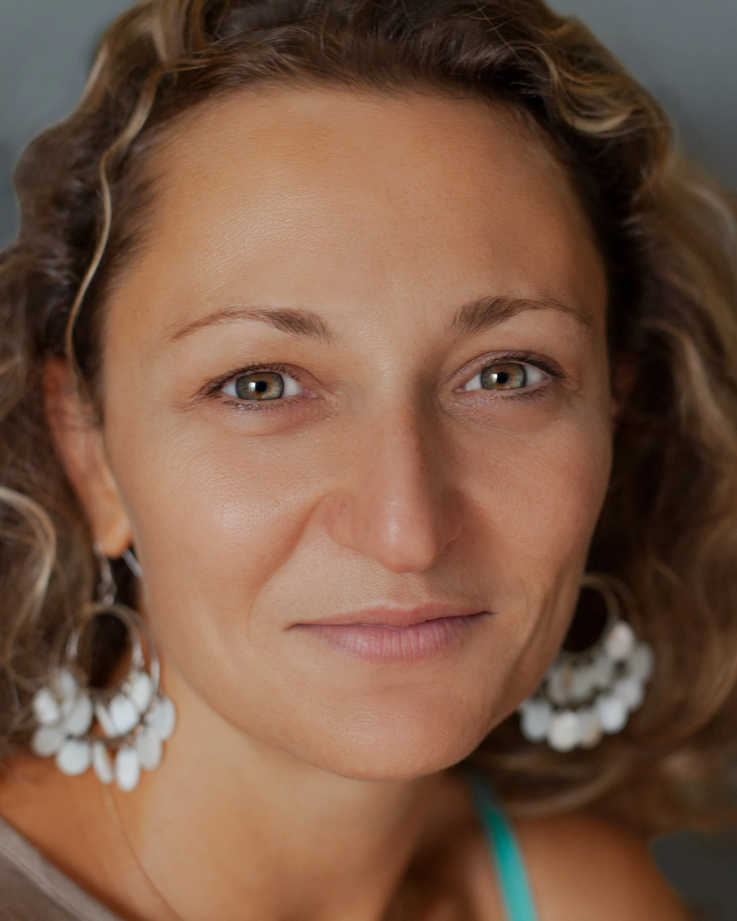 Close-up of a woman's face with curly hair, green eyes, and a slight smile, wearing white floral earrings and a teal strap.