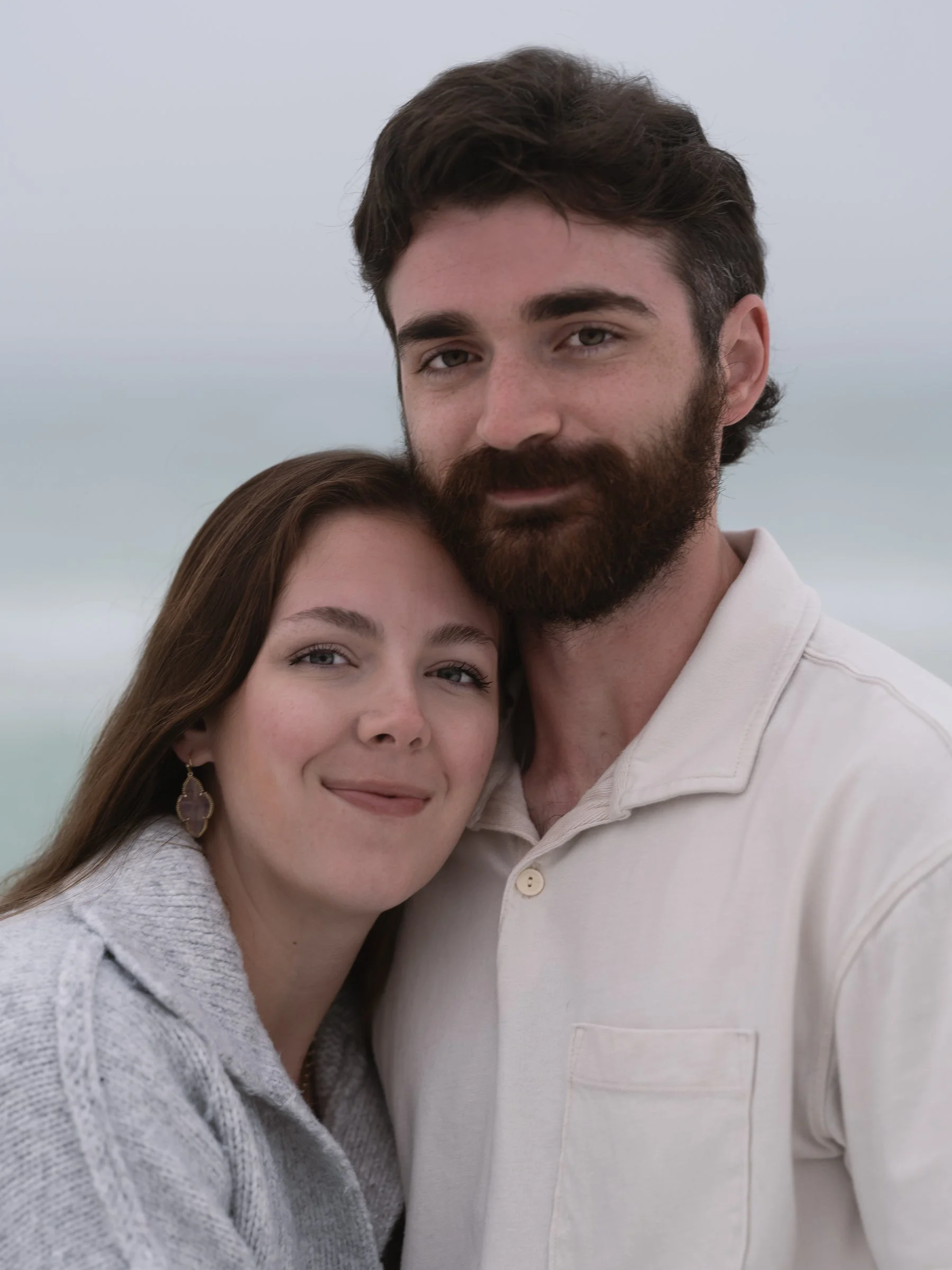 Close-up of a smiling woman with brown hair and earrings leaning her head on a smiling man with dark hair and a beard, both embracing each other outdoors.