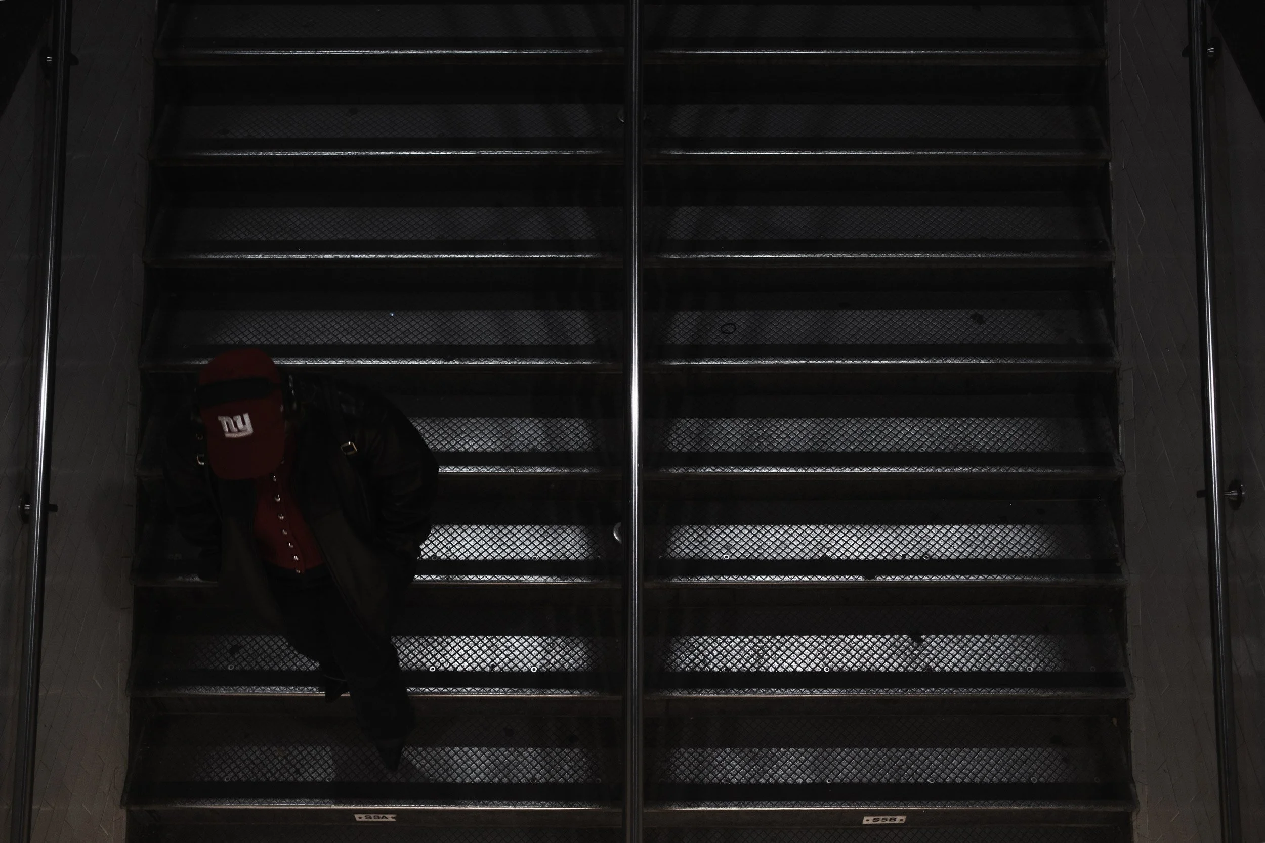 A person with a red and black backpack walking down a dark metal staircase, seen from above.