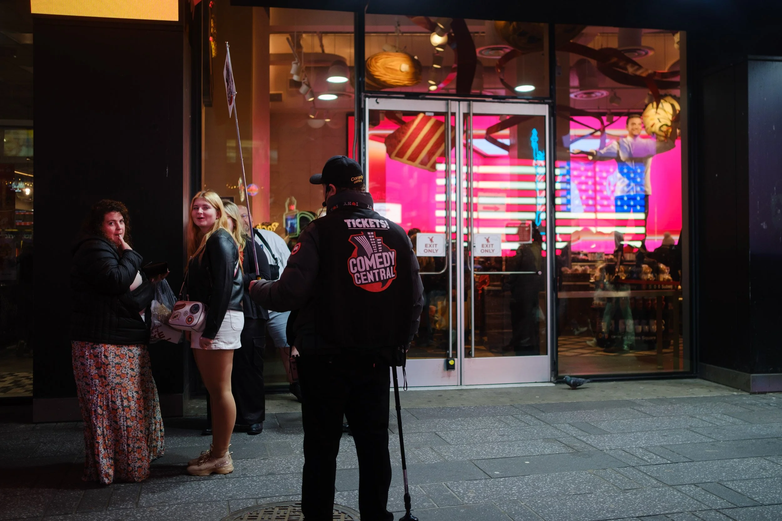 People standing outside a comedy club at night, with a clown in the background on a digital screen, and a performer preparing to entertain nearby.