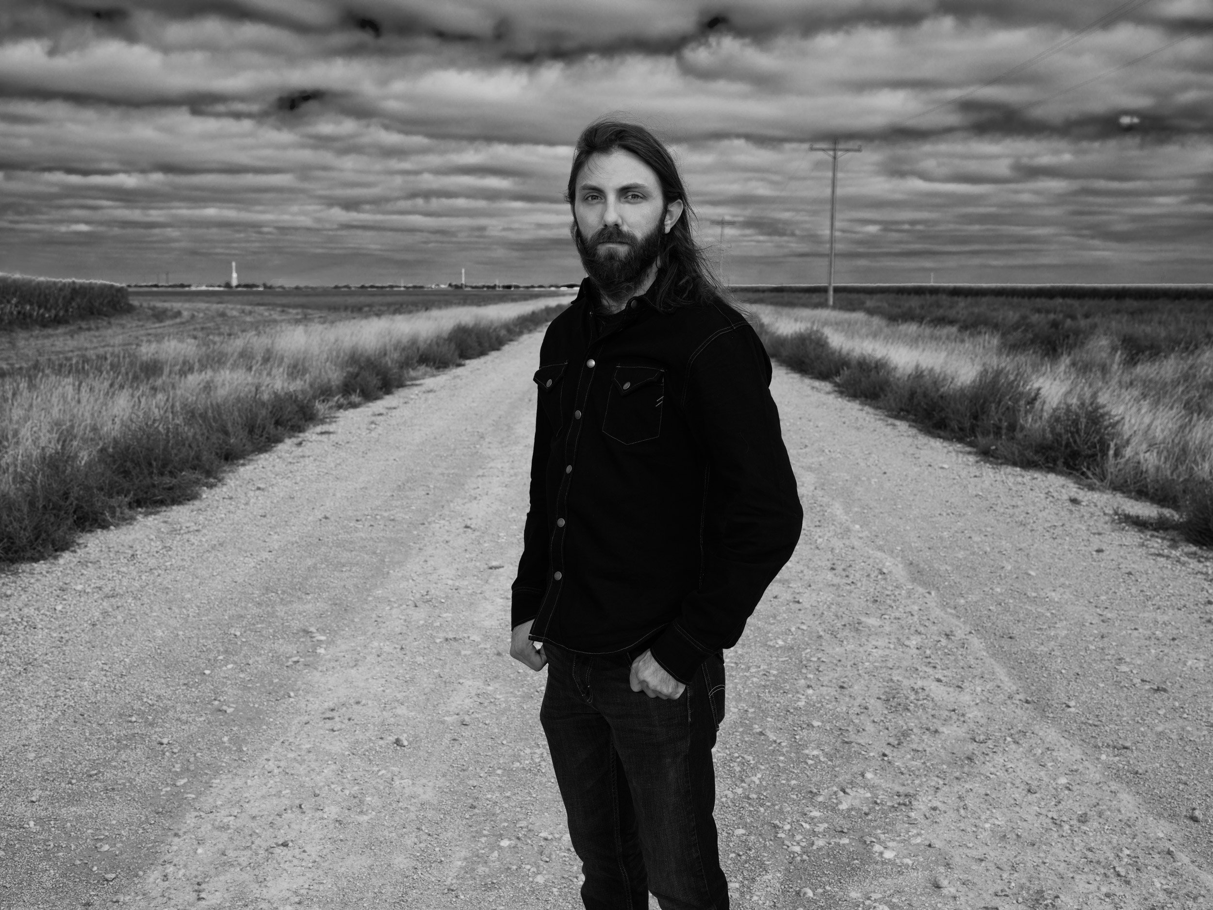 A man with long hair and a beard standing on a gravel dirt road in black and white, with open fields and a cloudy sky in the background.