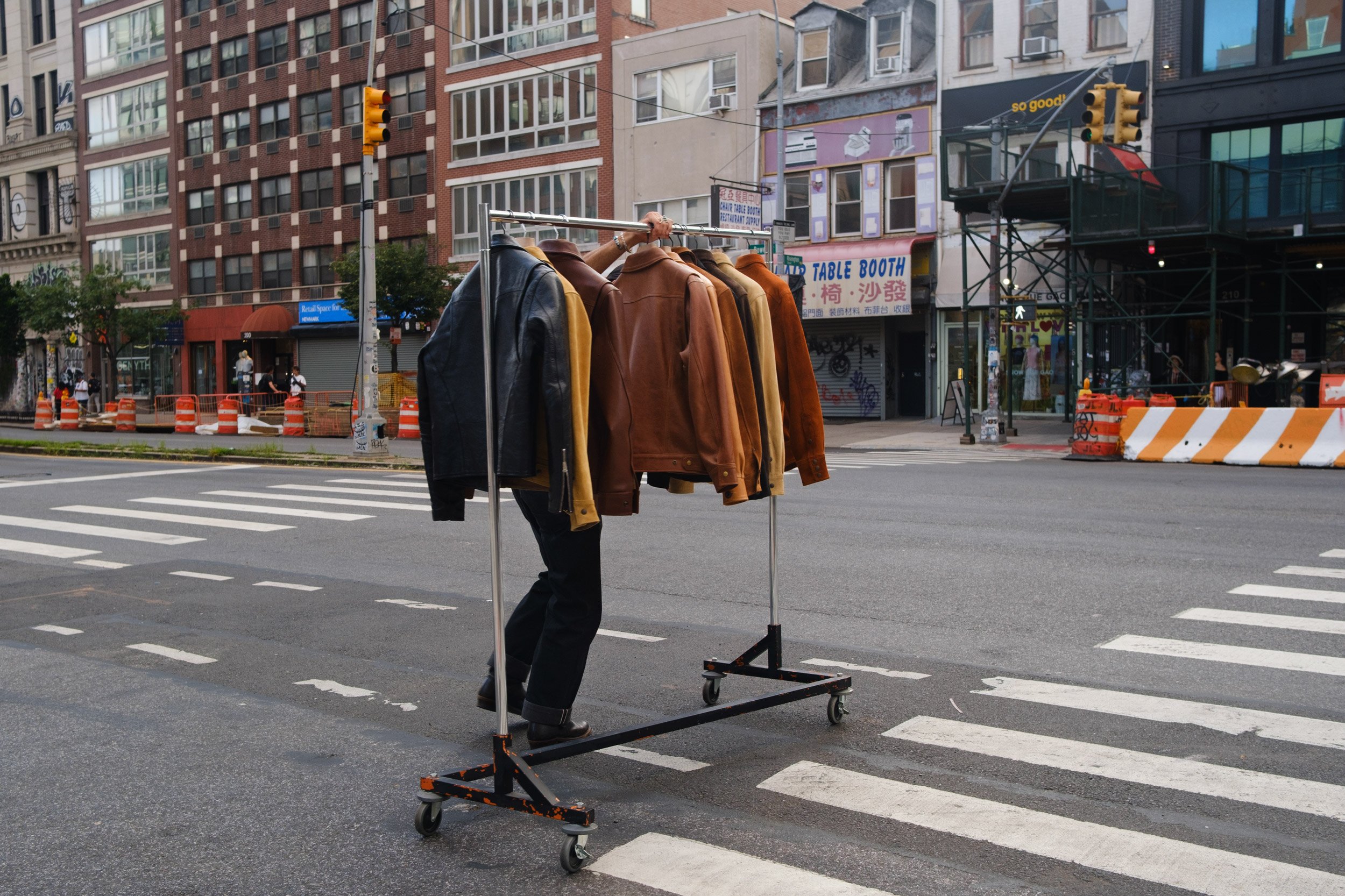 A man looking away, wearing a hat and dark clothing, holding a clothing rack with brown and black jackets in an urban street scene with multiple buildings, traffic lights, and construction barriers.