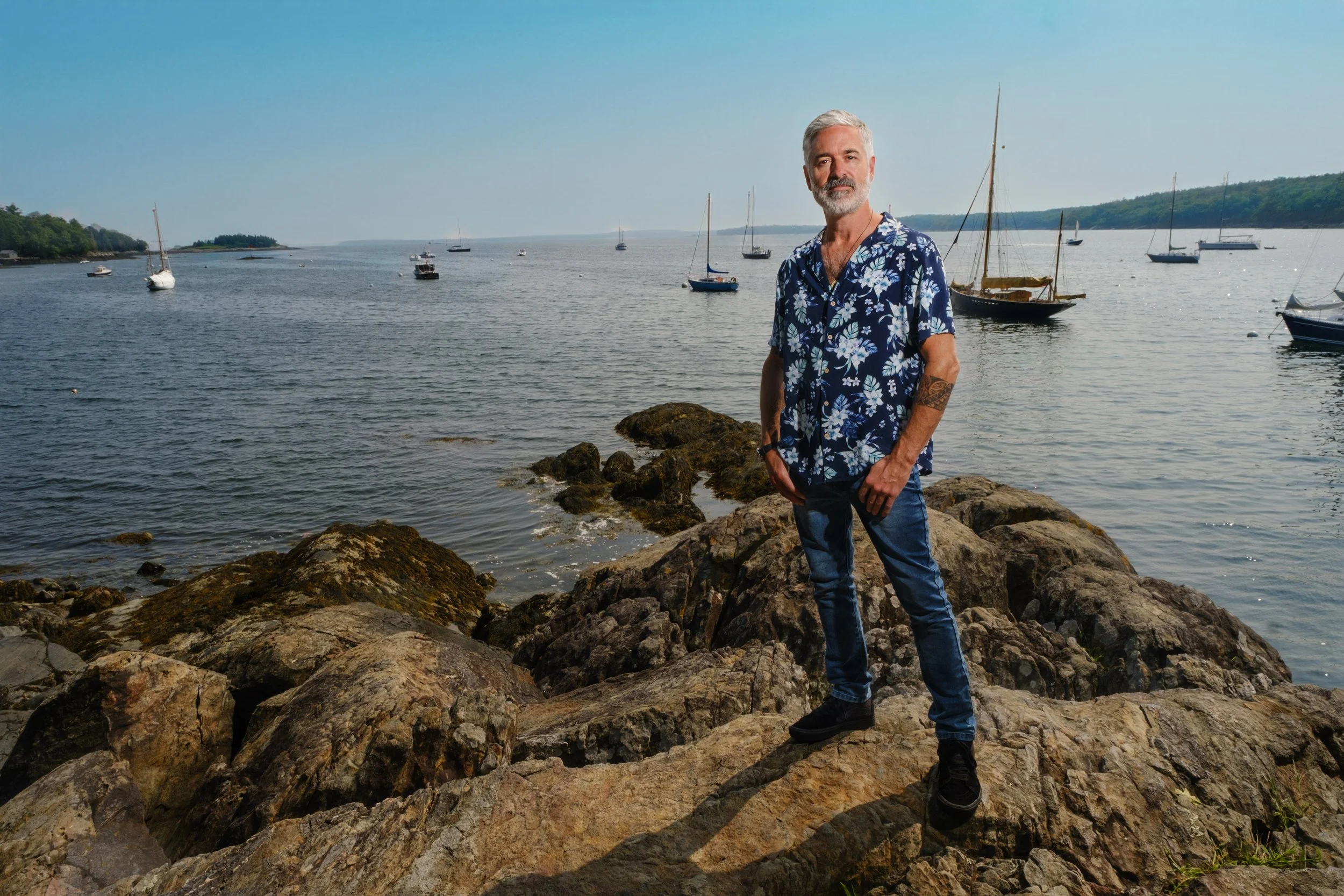 A man with gray hair and a beard standing on rocky shoreline near water with sailboats and hills in the background, wearing a blue Hawaiian shirt with white floral pattern and jeans.