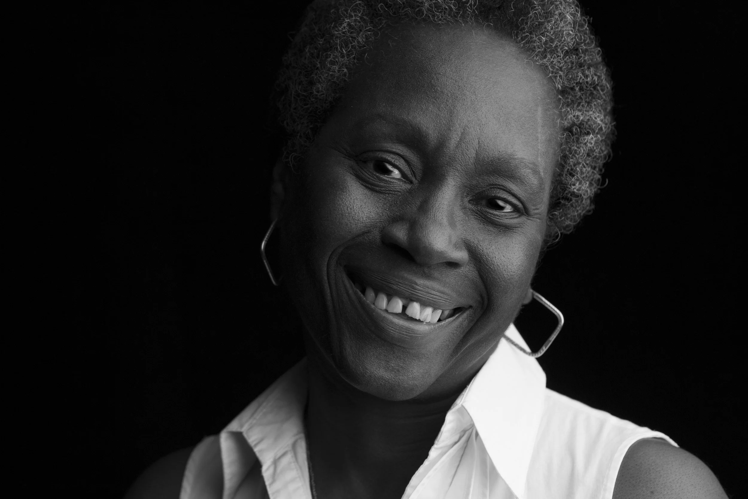 Close-up black and white photo of an older woman with short, curly, gray hair, smiling, wearing a white collared shirt, and hoop earrings.