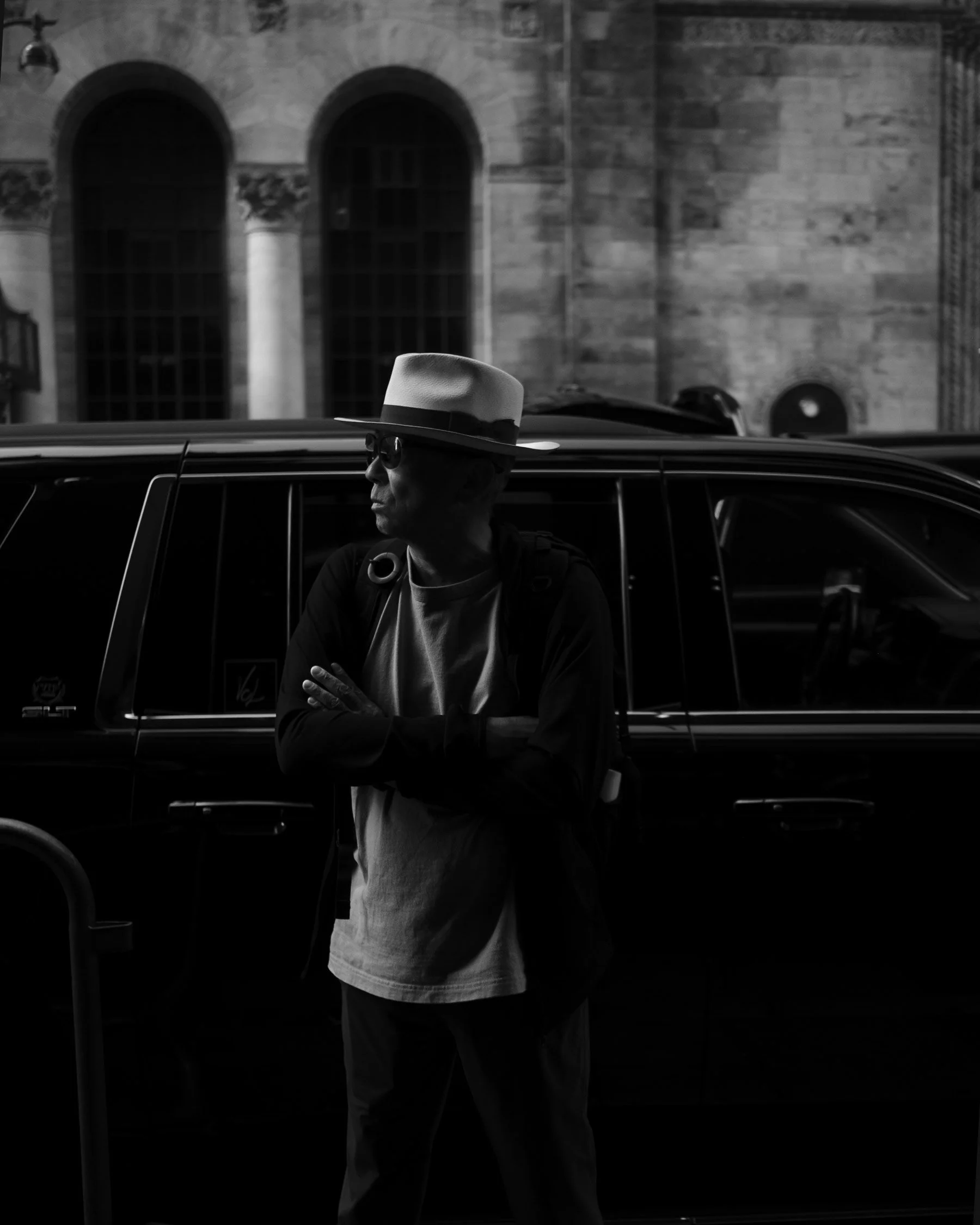 A man wearing a wide-brimmed hat and sunglasses standing outside a building with arched windows, leaning against a vehicle.