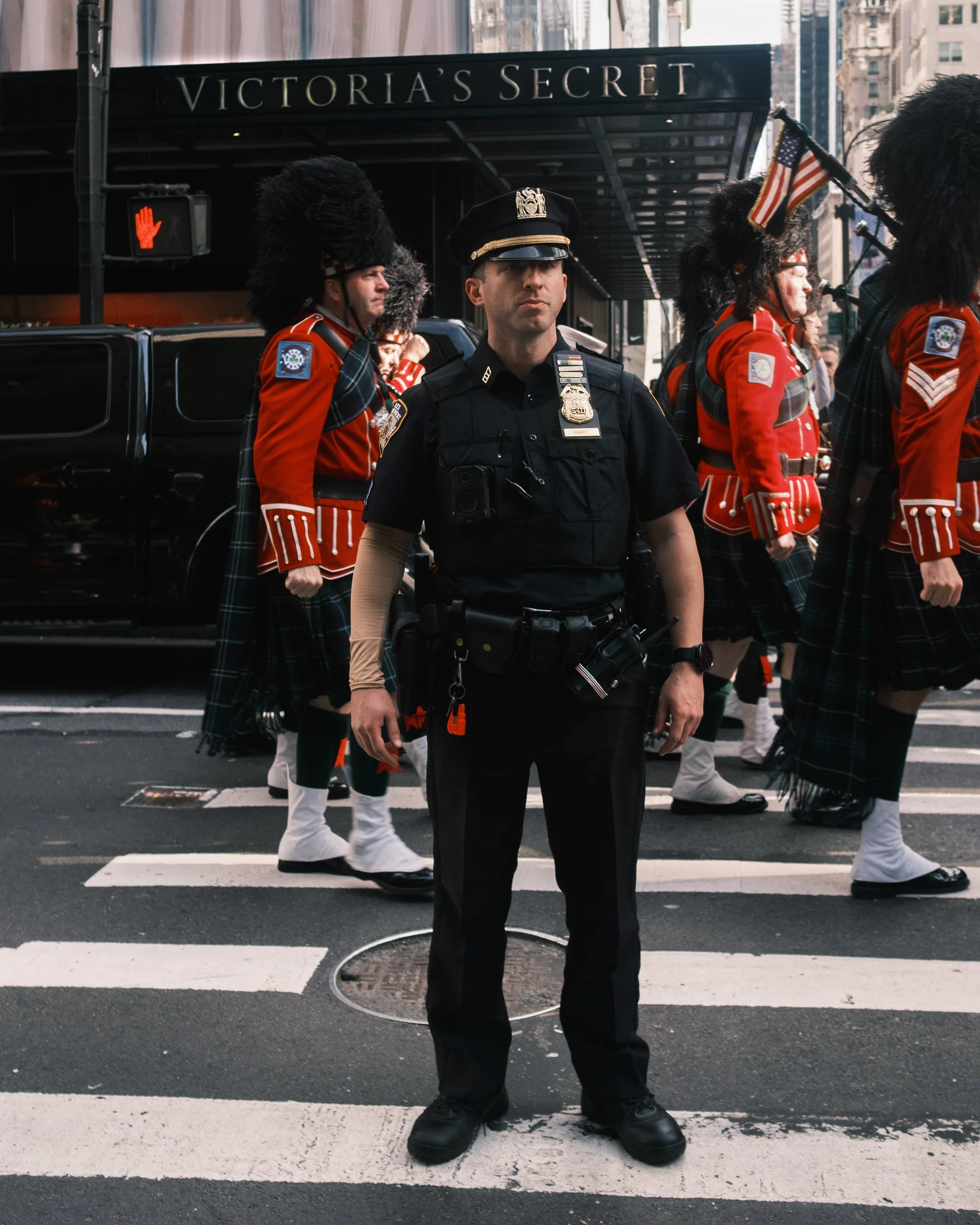 A police officer standing in the middle of a crosswalk on a city street, with members of a parade dressed in red uniforms and black hats with large feathers passing by. The background features a storefront with a sign that reads "Victoria's Secret" a