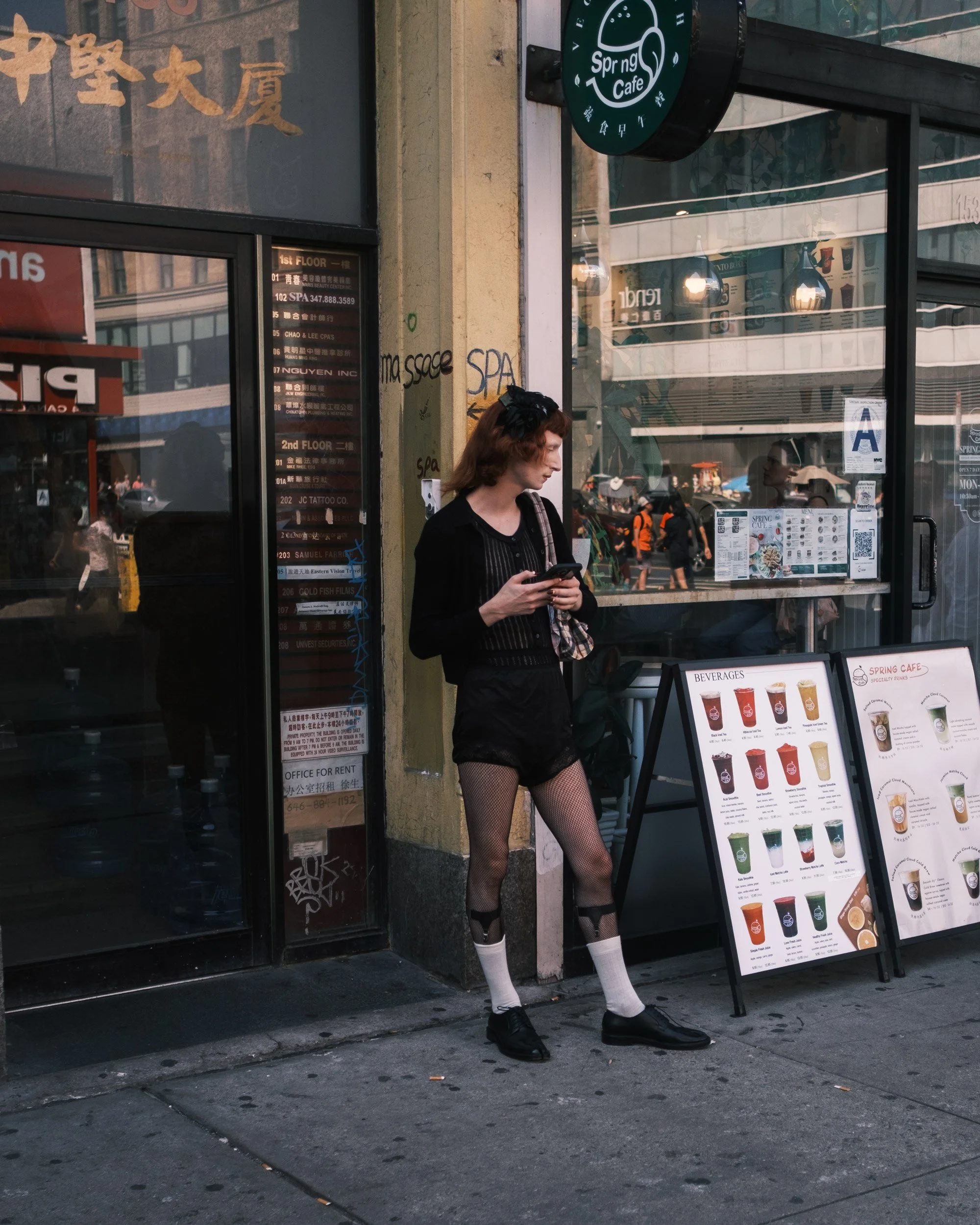 A person with red hair dressed in black fishnet stockings, a black top, and shoes, standing outside a cafe while looking at their phone. The person is wearing a black headband. There is a menu board outside the cafe displaying various beverages. The 