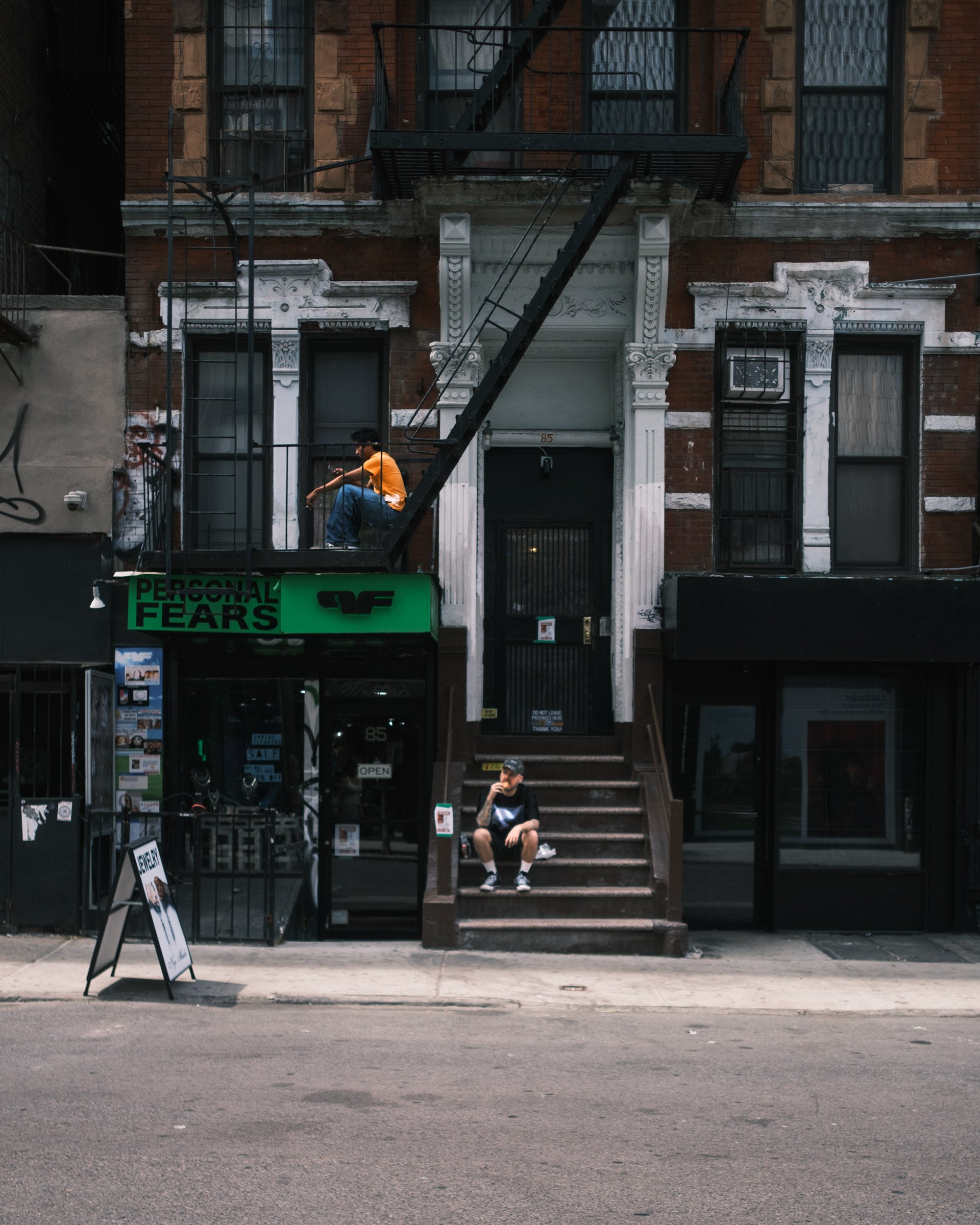 A city street scene with a brownstone building featuring fire escapes. Two young men are sitting on the fire escape steps, one wearing an orange shirt and jeans, the other in a black shirt and shorts. The street has storefronts at ground level, one w