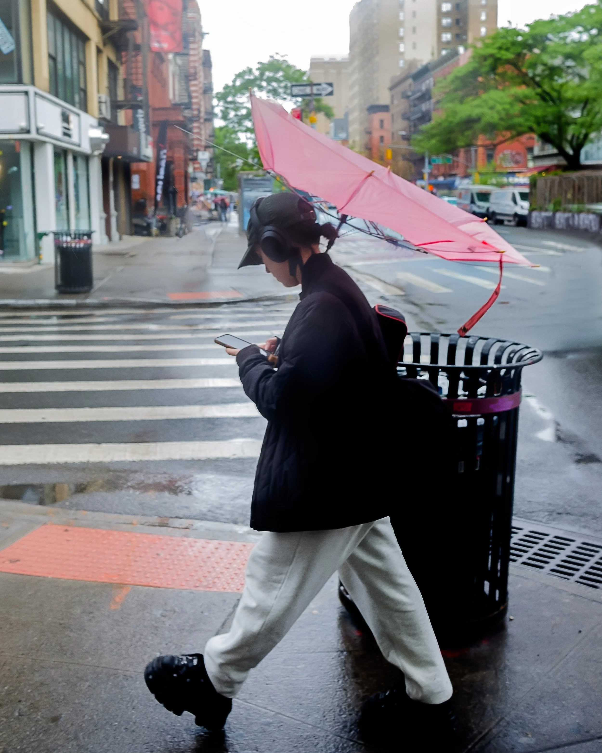 A person walking on a city street holding a pink umbrella and looking at a smartphone. The person is wearing a black hoodie, beige pants, and black shoes. The street is wet with puddles, and buildings, trees, and parked cars are visible in the backgr