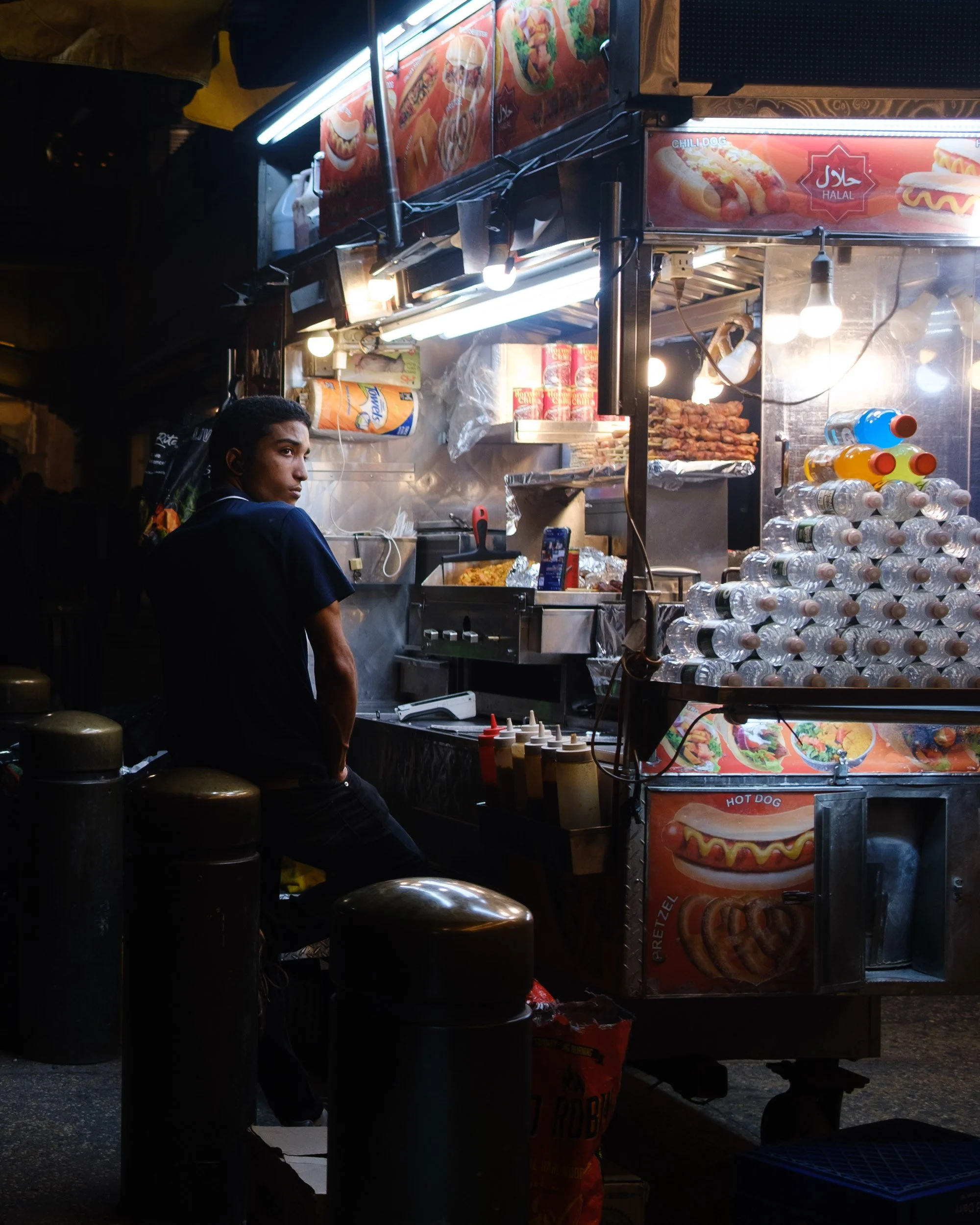 Street food stall at night selling hot dogs, buns, and drinks, with a person standing nearby.