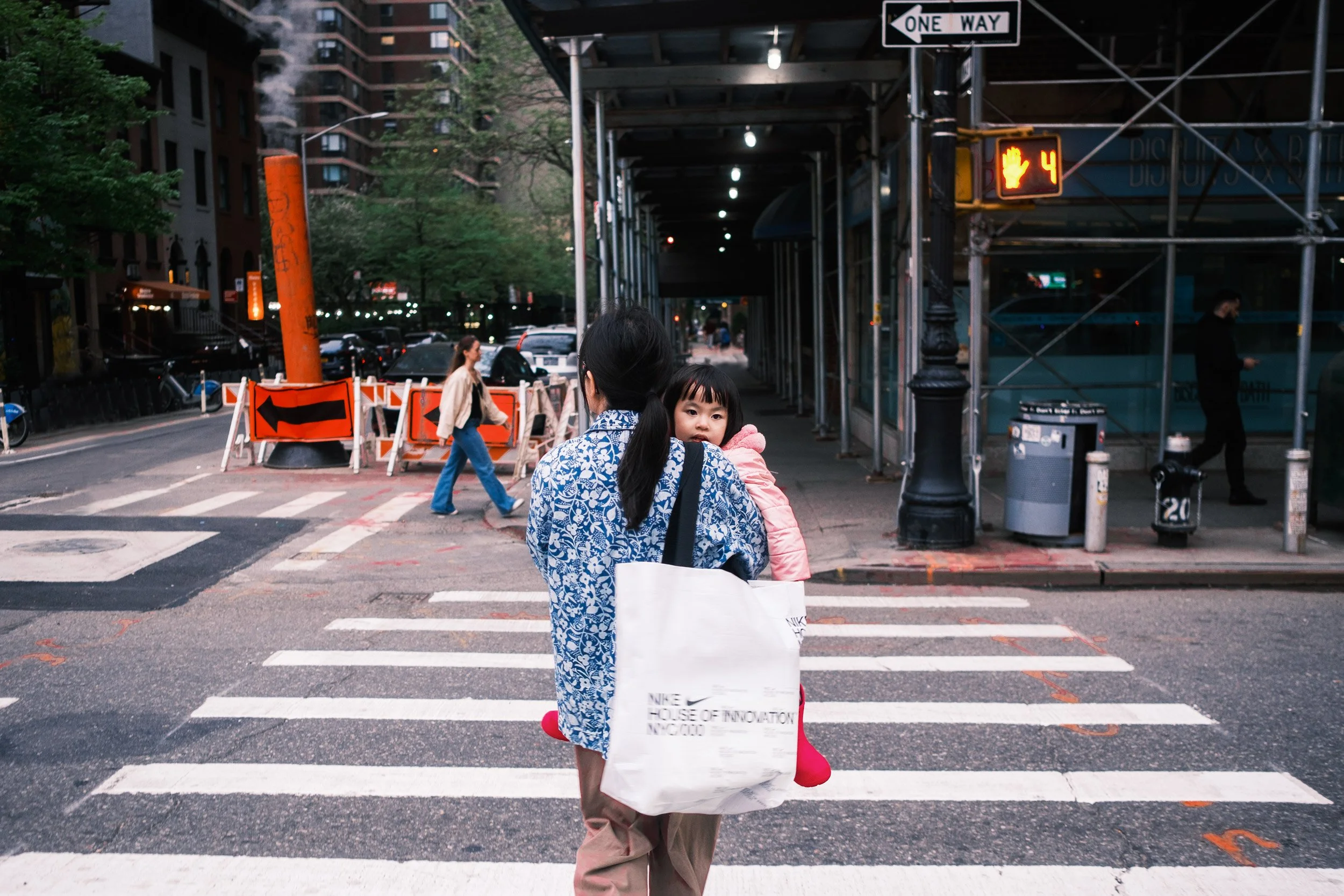 A woman carrying a child crossing the street on a crosswalk in an urban area. There is a woman walking on the sidewalk, construction barriers, and signs including a one-way sign, a pedestrian signal showing a hand with the number 4, and a stop sign. 
