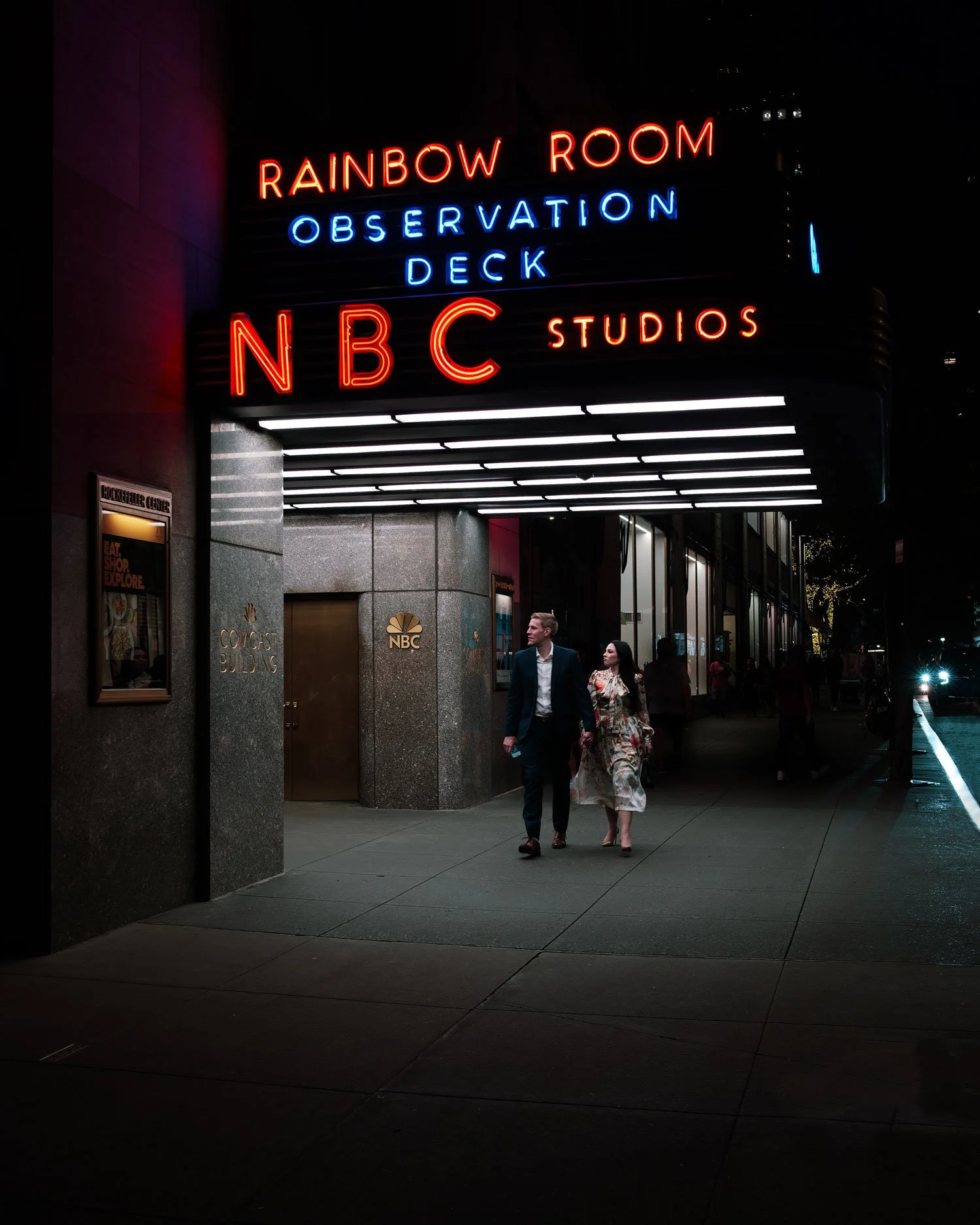 Night view of the NBC Studios building with illuminated sign reading "Rainbow Room Observation Deck NBC Studios" in neon lights. People walking outside the building.
