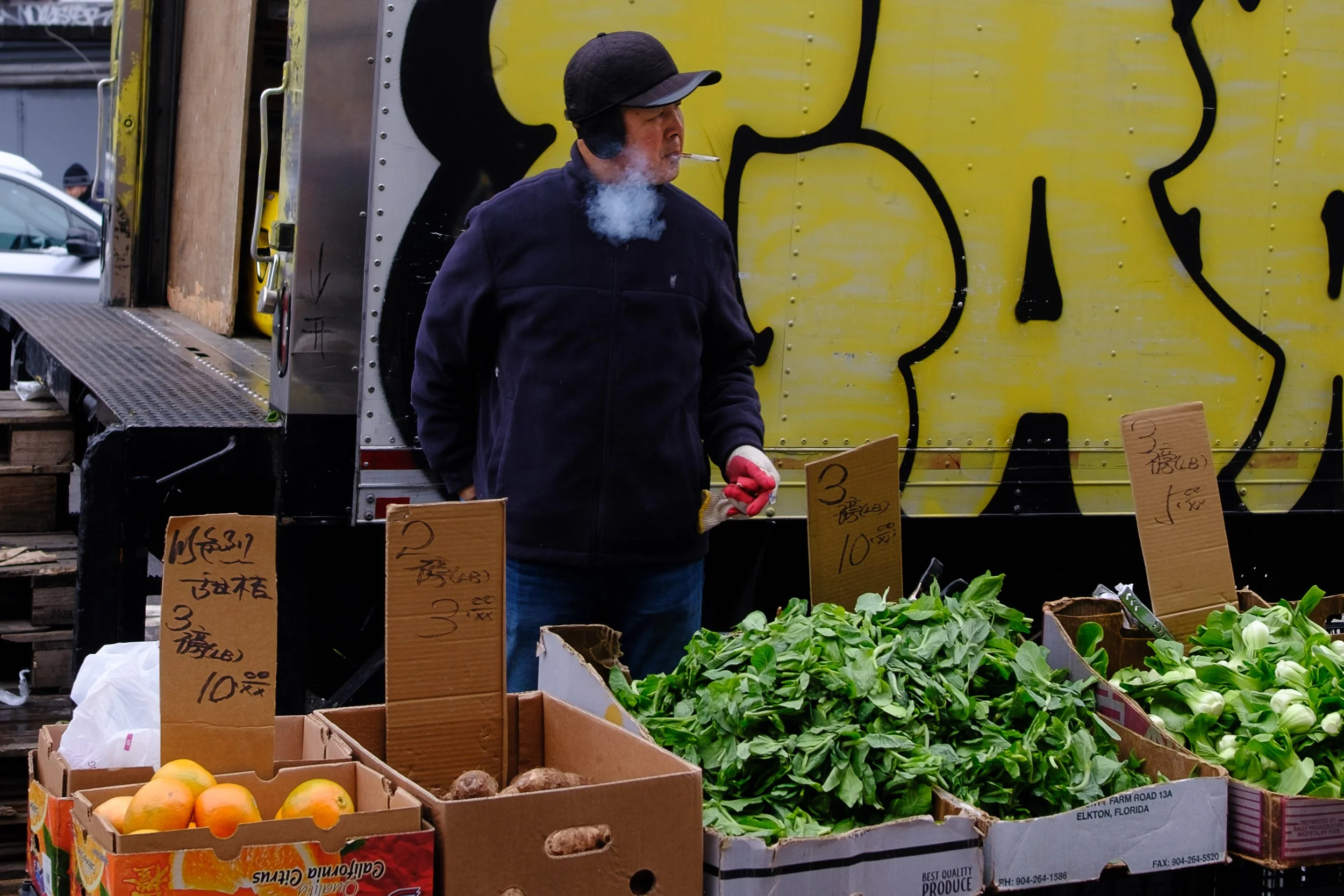 A man smoking a cigarette at a fruit stand with boxes of apples, vegetables, and leafy greens, and handwritten price signs. He is wearing a black jacket and a black helmet.
