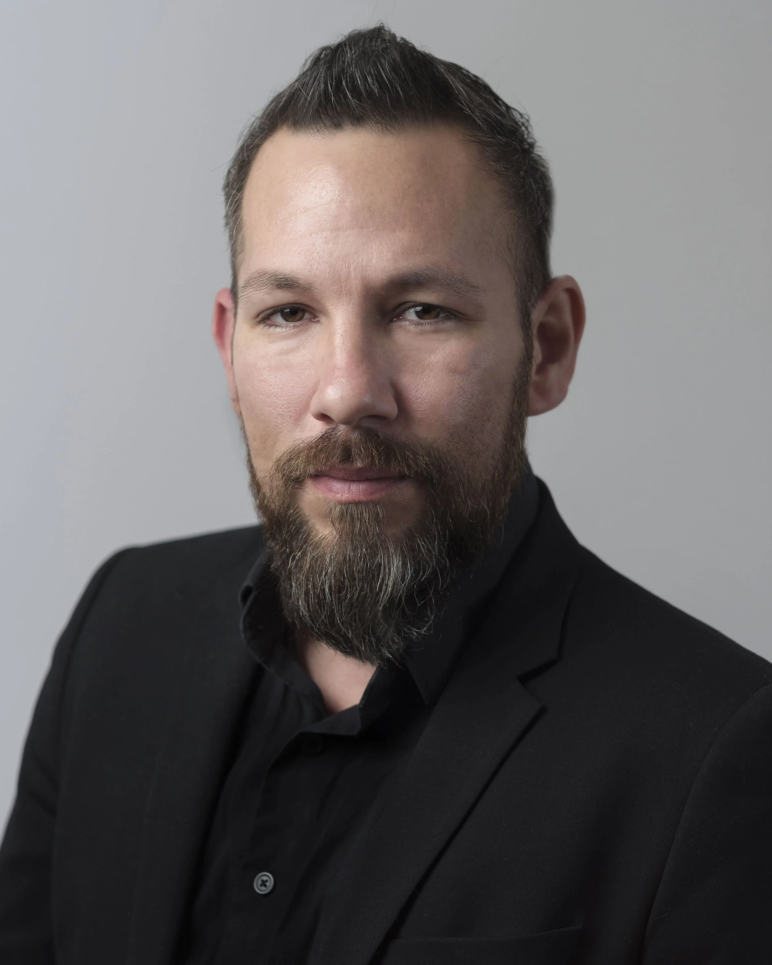 Close-up of a man wearing a black suit with a collared shirt, with short dark hair styled upwards and a full beard, against a light gray background.