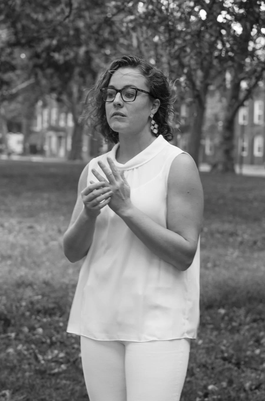 Black and white photo of a woman with glasses and curly hair, wearing a sleeveless white top and white pants, standing outdoors with trees and buildings in the background. She appears contemplative or emotional, with her hands clasped near her chest.