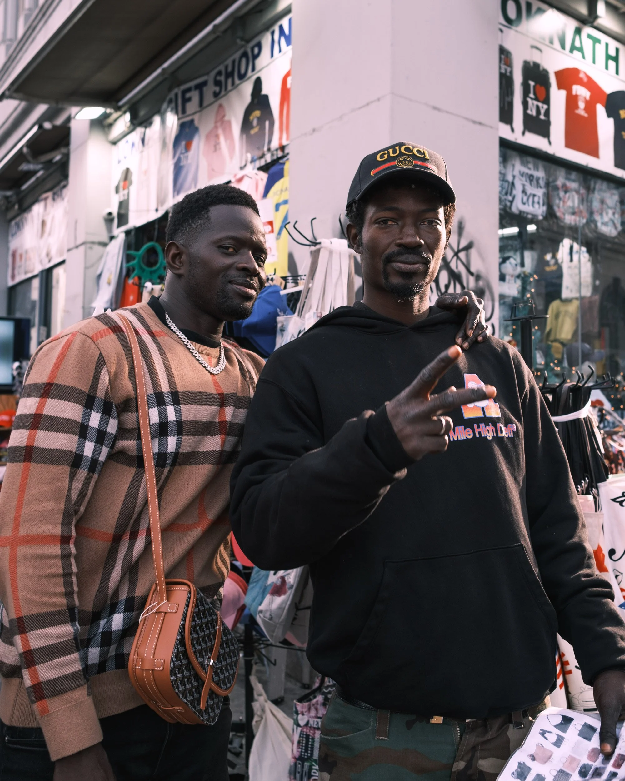 Two men standing in front of a street vendor stall filled with clothing and accessories. One man wears a checkered brown and beige jacket with a silver chain necklace and carries a tan crossbody bag. The other man wears a black Gucci cap and a black 