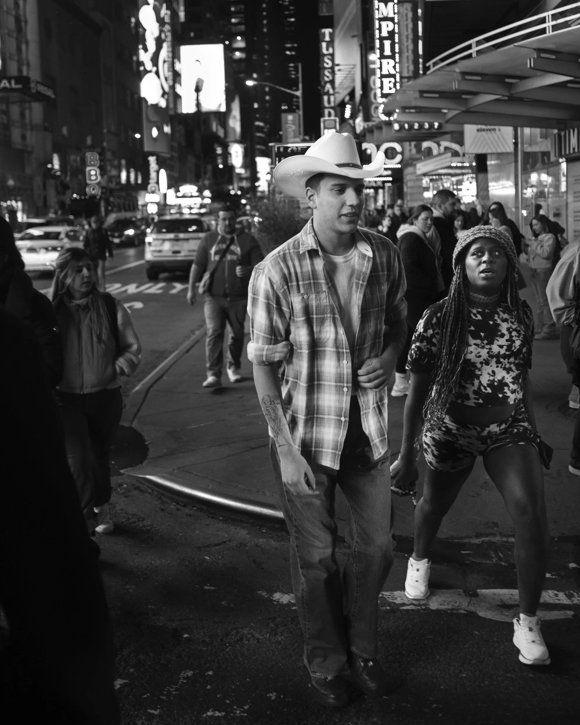 Black and white photo of people walking on a city street at night, illuminated by bright city lights and signs, with a young man wearing a cowboy hat and a woman with long braided hair in the foreground.