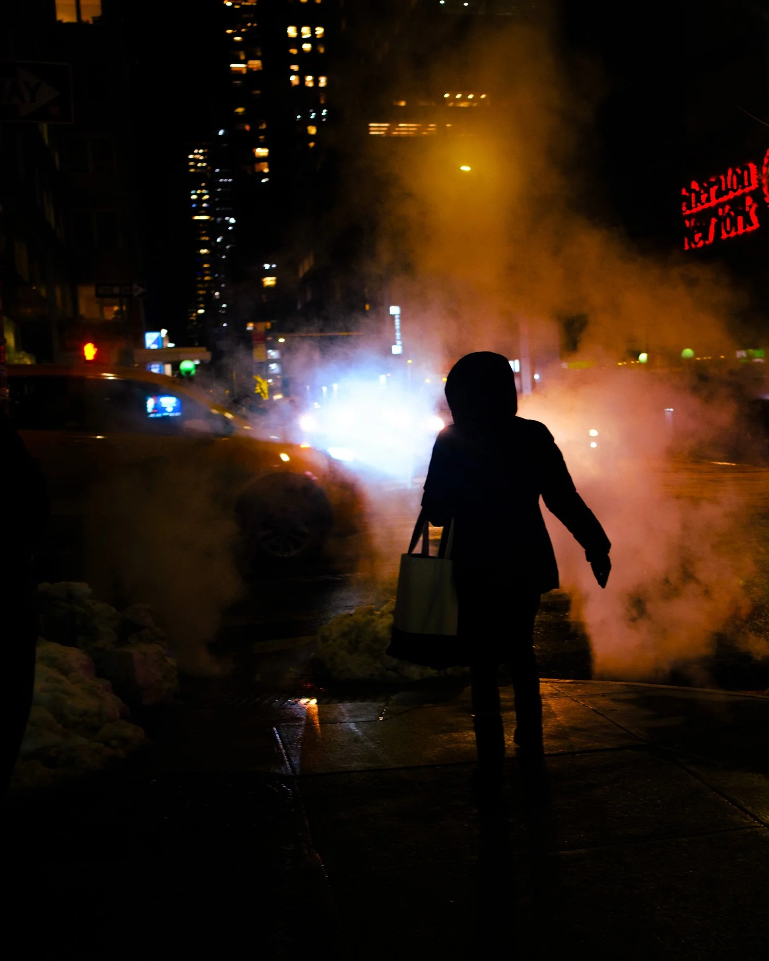 A person wearing dark clothing and a hood walking on a wet sidewalk at night, with steam or smoke rising in the background, city lights and buildings visible in the distance.